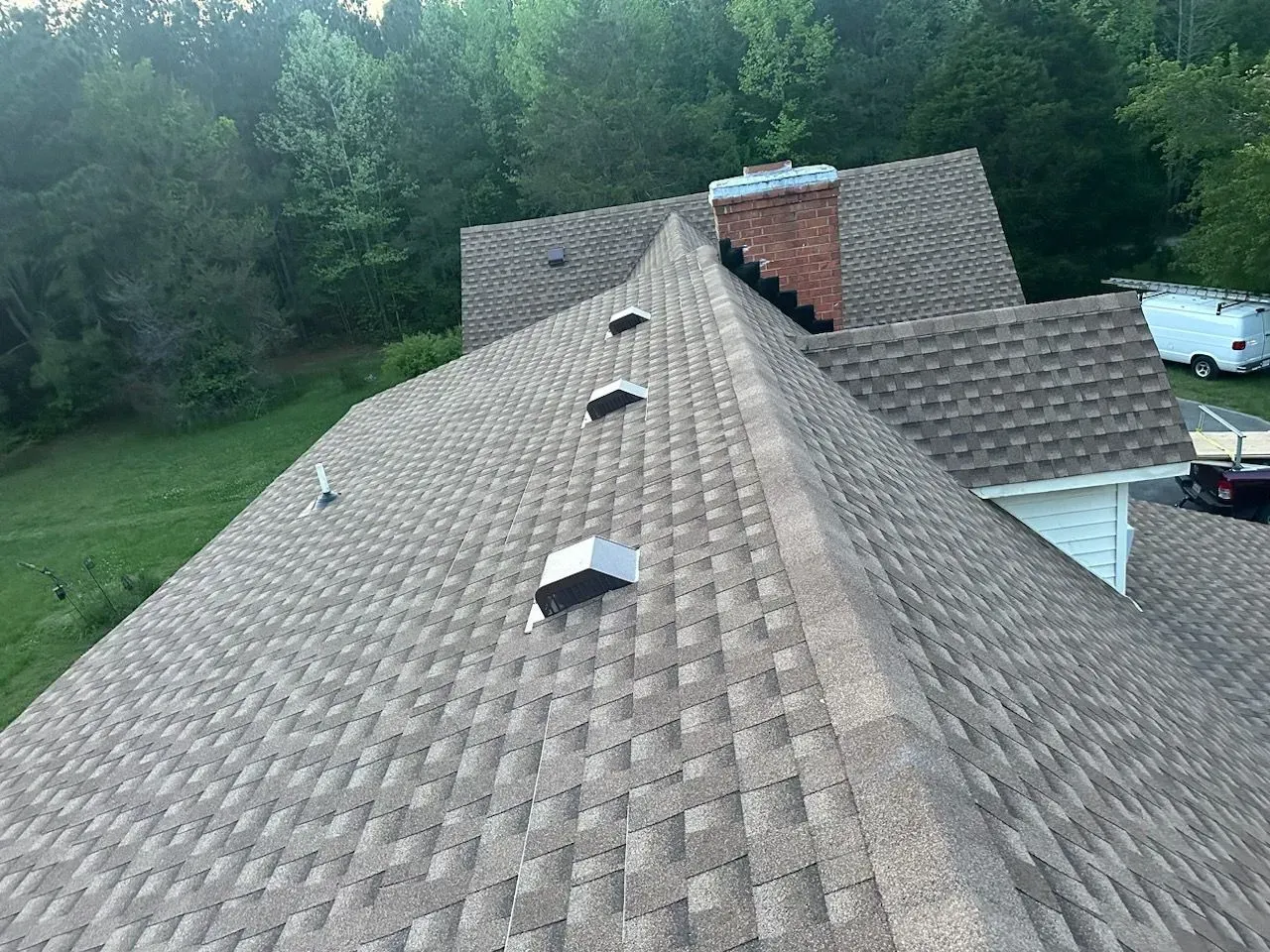 Overhead view of a house roof with brown shingles, vents, and a brick chimney against a green tree backdrop.