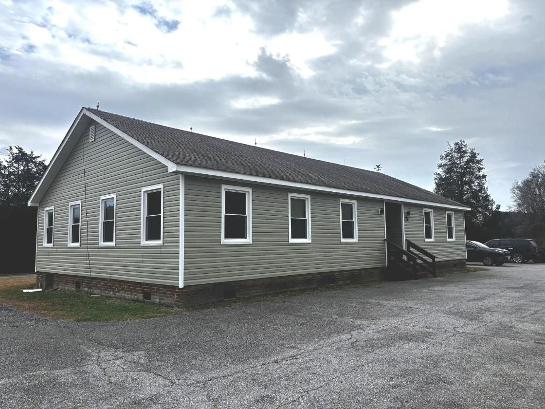 Light gray building with multiple windows under a cloudy sky, gravel parking area.