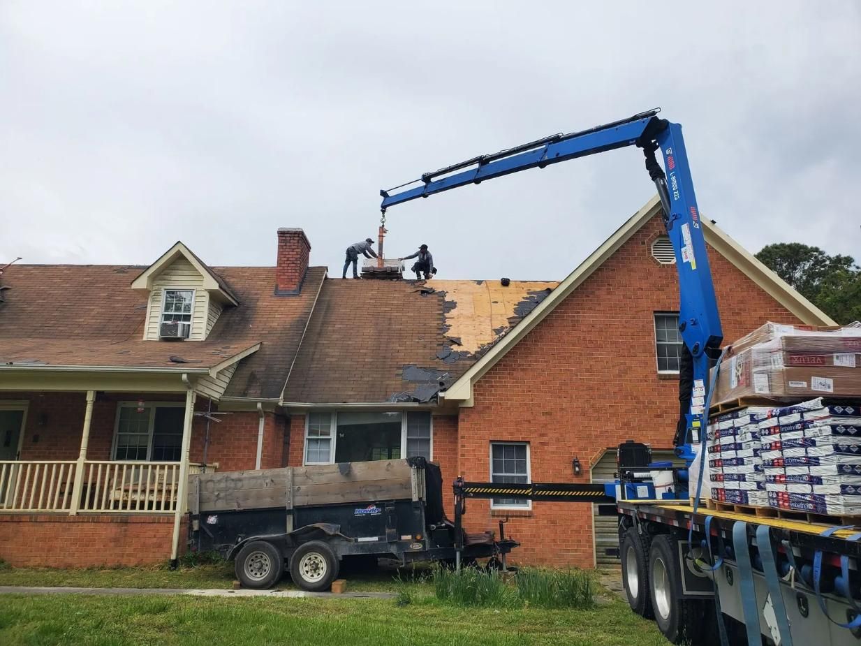 Roofers using a crane to install shingles on a brick house with a trailer and a flatbed truck on a cloudy day.