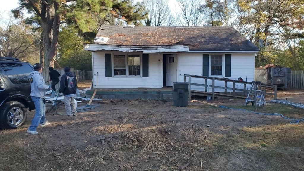 A small, weathered white house with a damaged roof and a wooden ramp. 