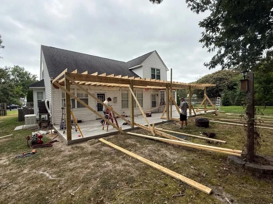 Construction workers building a wooden patio roof attached to a two-story white house on a cloudy day.