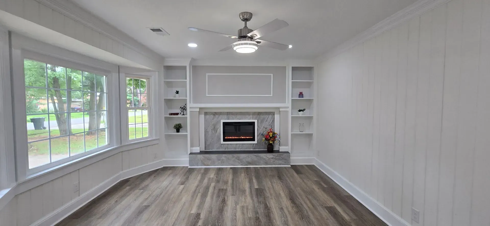 Living room with fireplace, built-in shelves, large window, wood flooring, and ceiling fan.