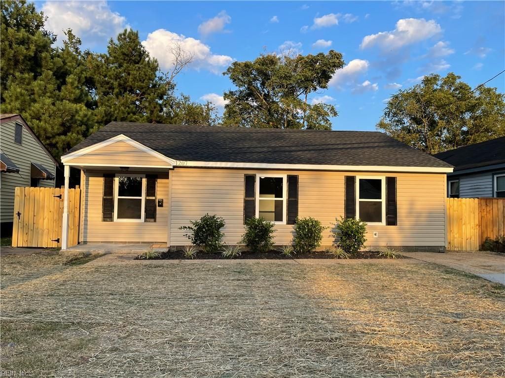 Tan bungalow with black shutters, small bushes, and brown fence.
