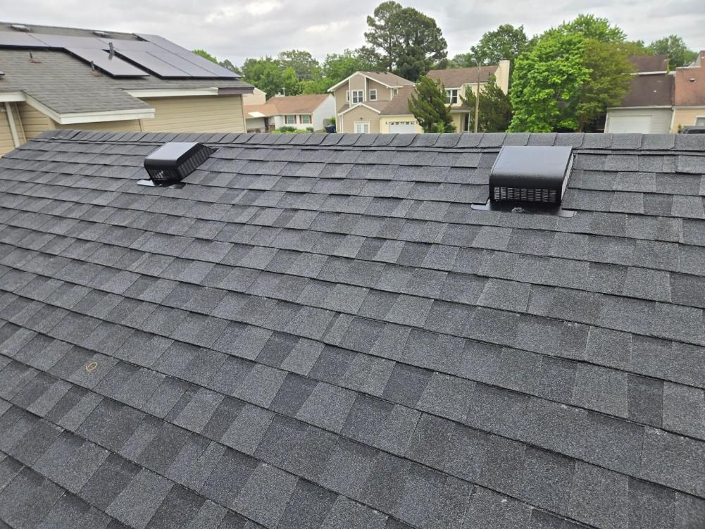 Dark shingle roof with two black vents. Houses and trees in the background under cloudy sky.