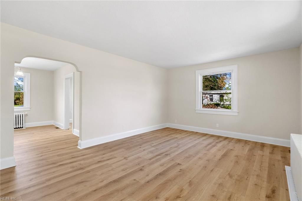 Empty living room with wood flooring, white walls, and a window. An arched doorway leads to another room.