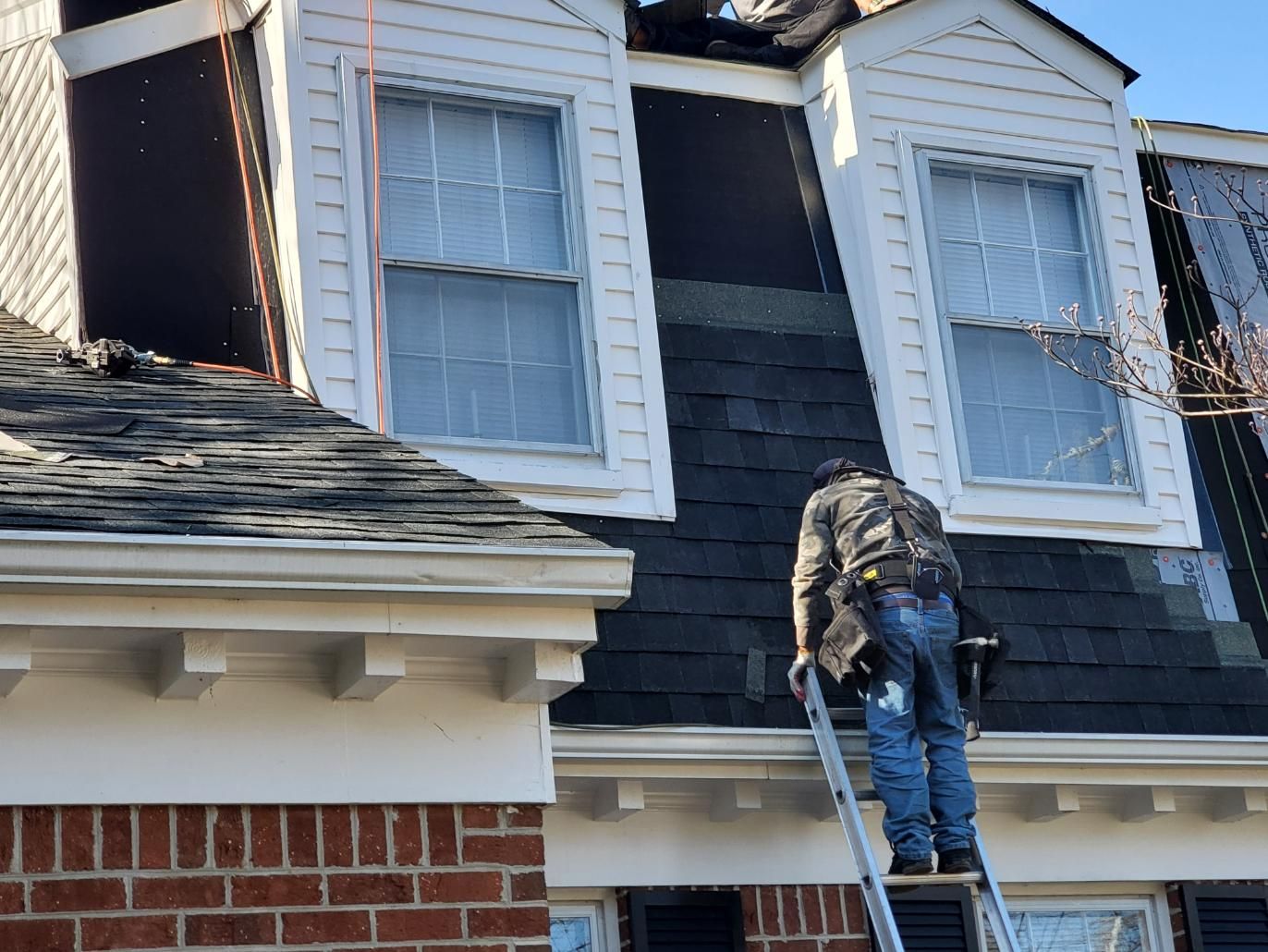 Roofer on a ladder installing shingles on a house roof with dormer windows.