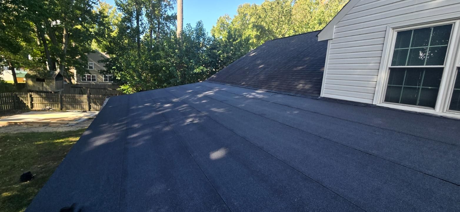 View of a house roof, covered in dark asphalt shingles, with trees and a clear blue sky in the background.