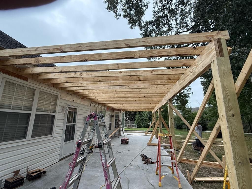 Pergola under construction, attached to a white house with a concrete patio.