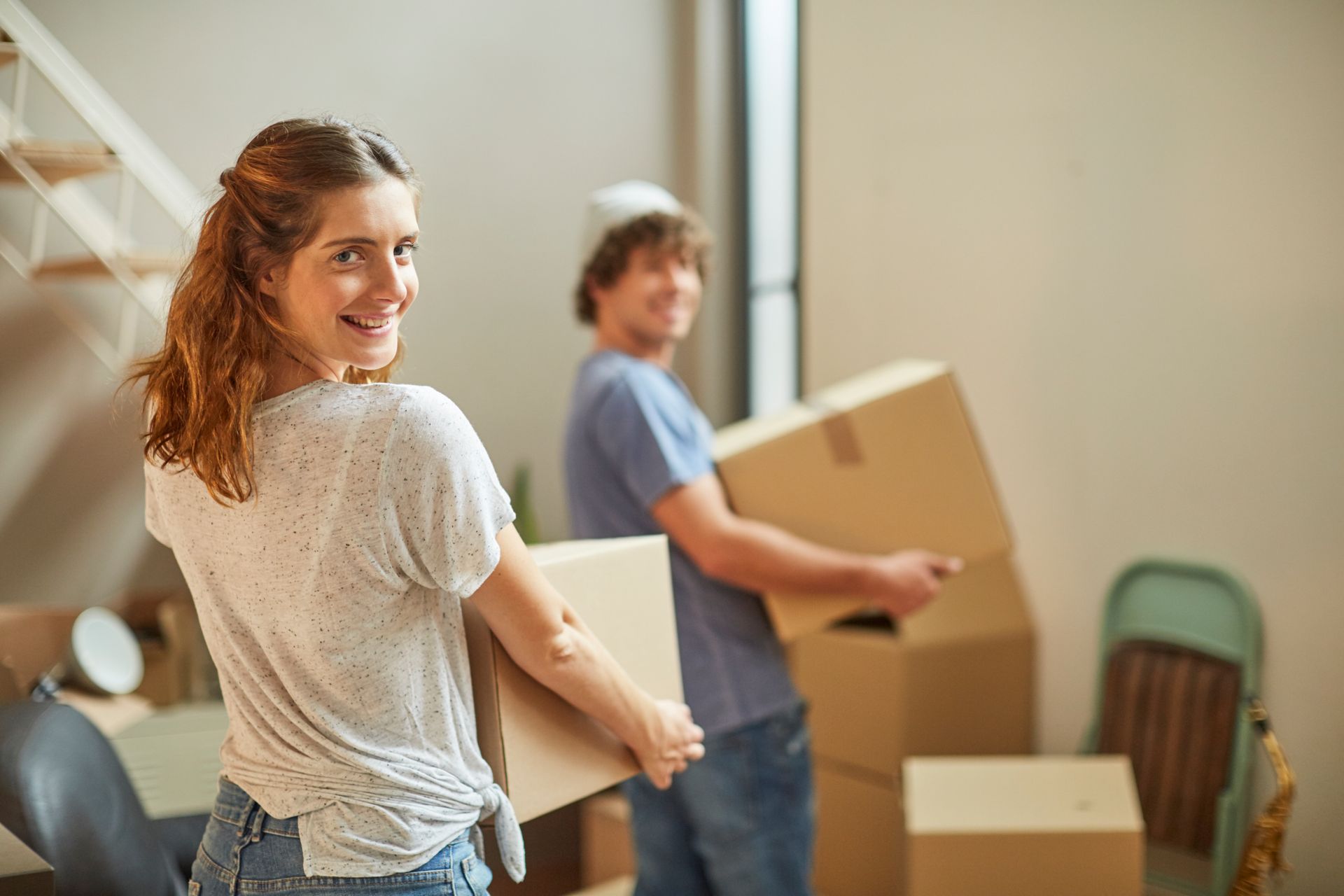 Woman and man smiling, carrying cardboard boxes in a light-filled room, possibly moving.