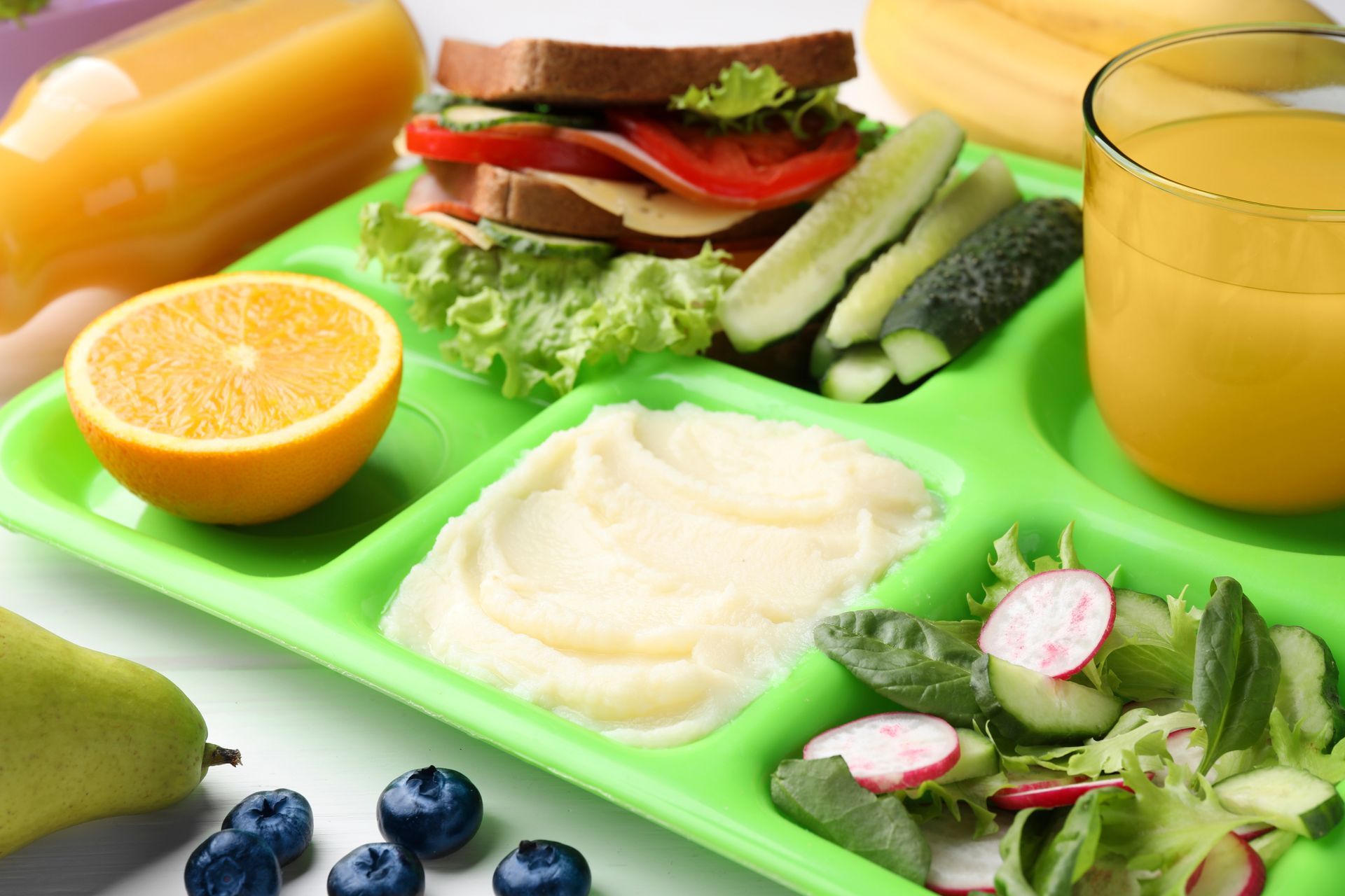 A green tray filled with sandwiches , vegetables , fruit and a glass of orange juice.