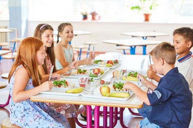 A group of children are sitting at a table in a cafeteria eating food.