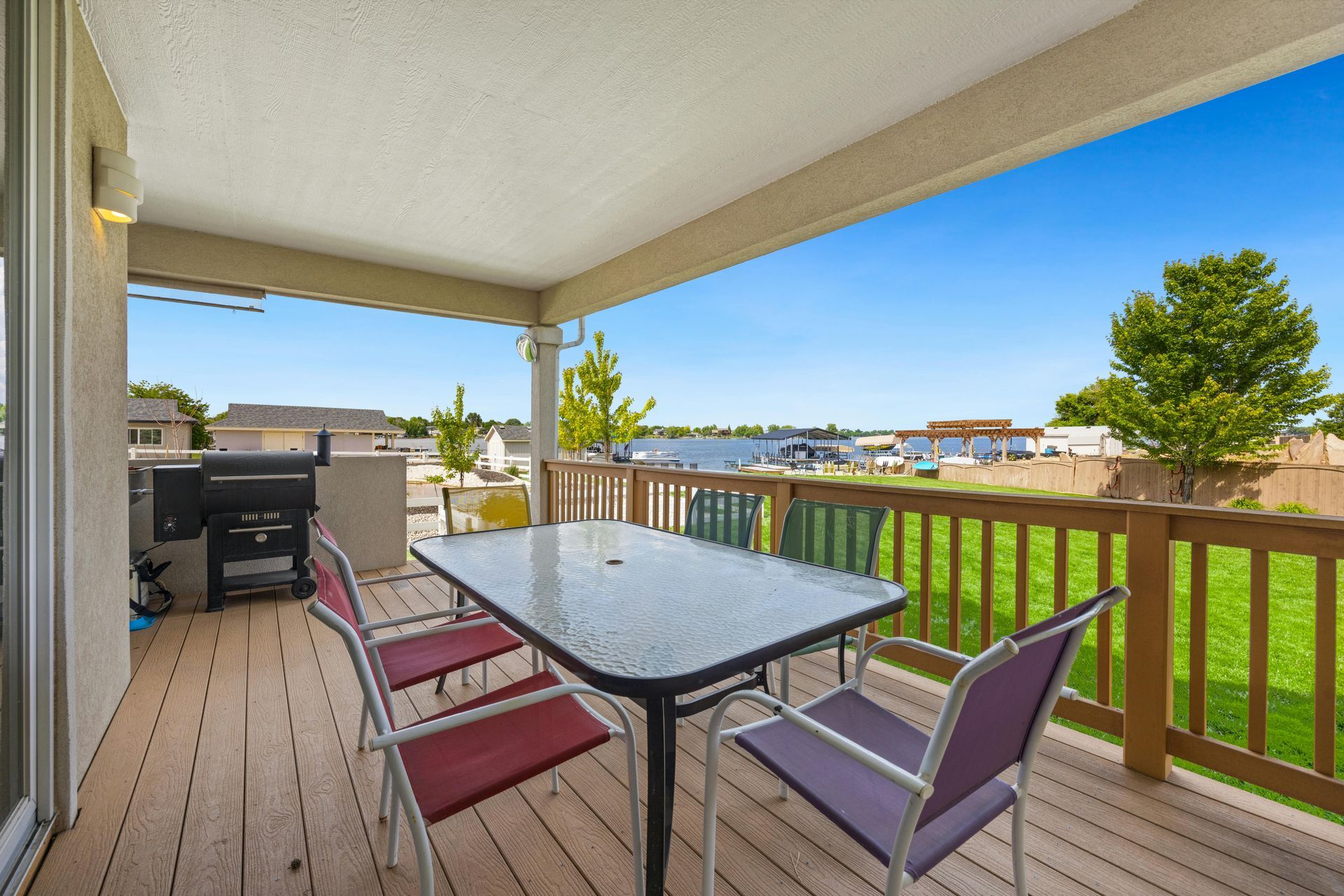 Patio with table, chairs, grill, overlooking a green yard and lake under a bright blue sky.