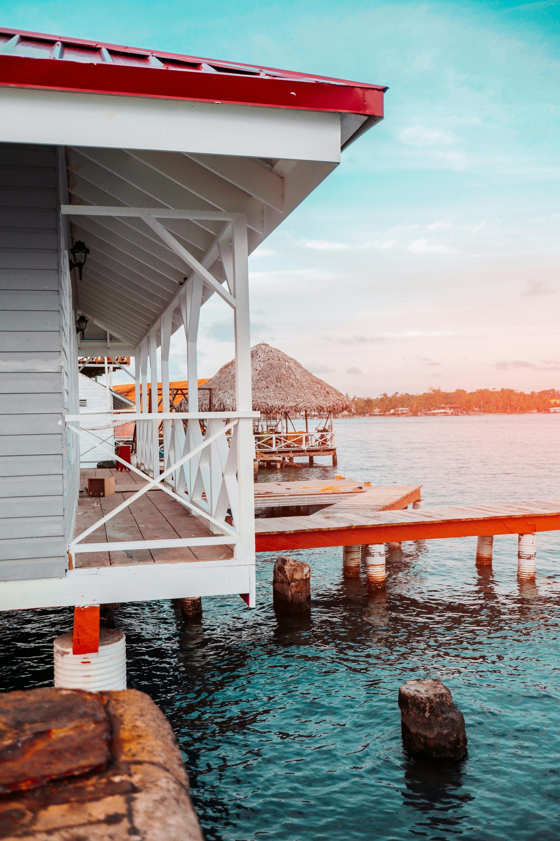 White stilt house with red trim over turquoise water, pier, and a thatched hut; bright sky.