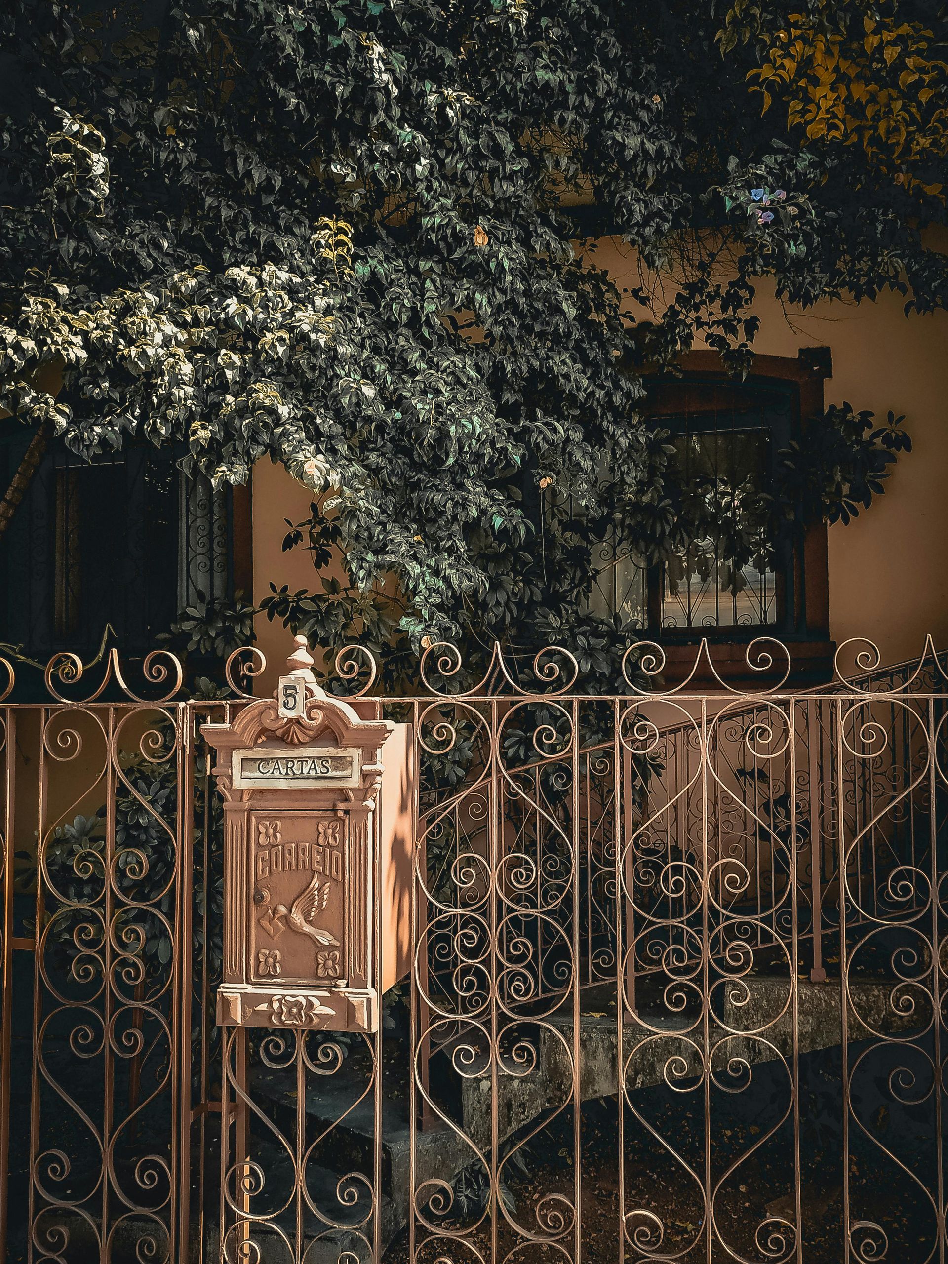 Ornate bronze mailbox on a matching wrought iron fence; a building and lush green tree in the background.