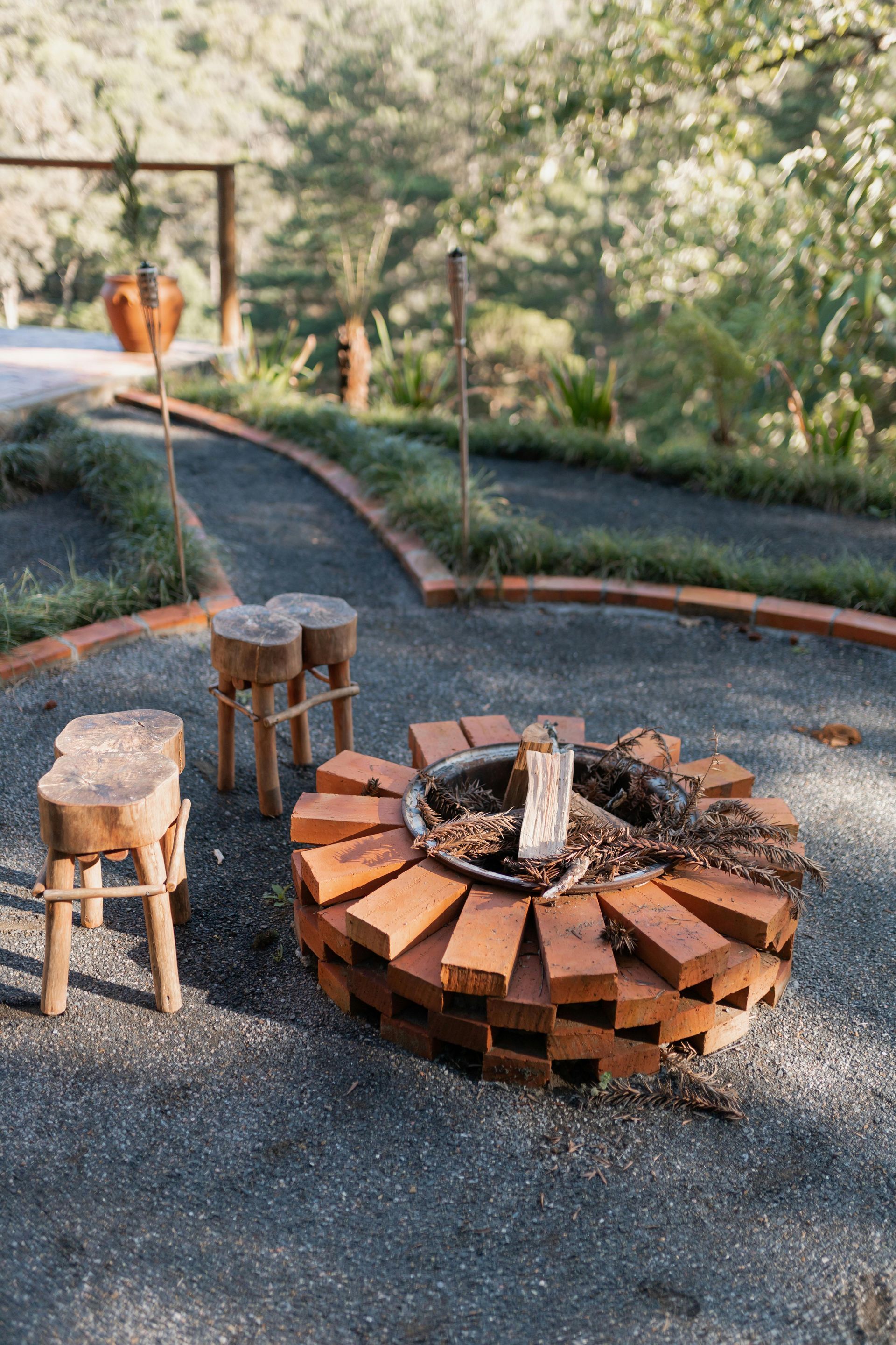 Brick fire pit with log stools on gravel, surrounded by a landscaped path and greenery.