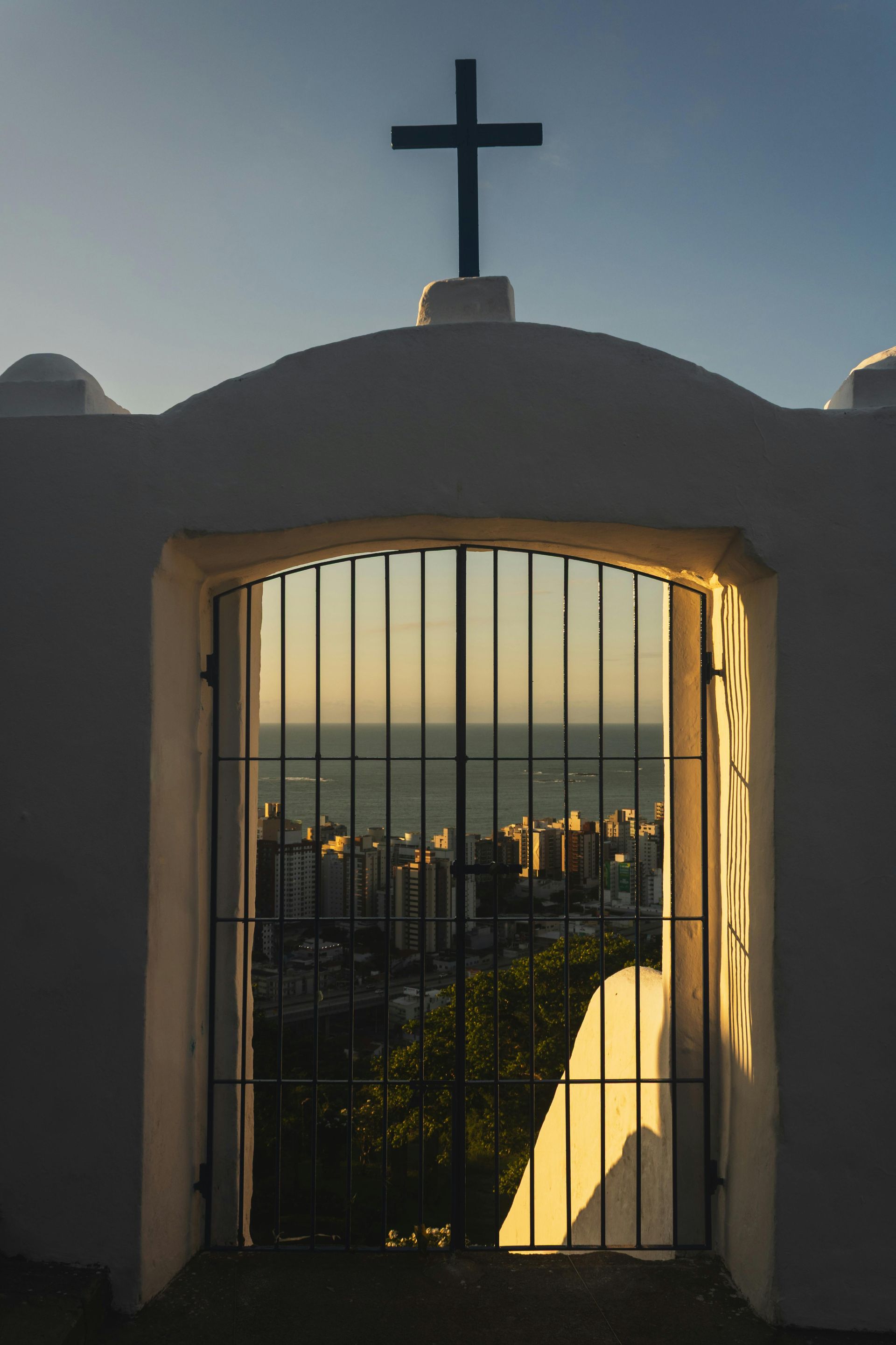 Arched white structure with cross, metal gate revealing city and ocean at sunset.
