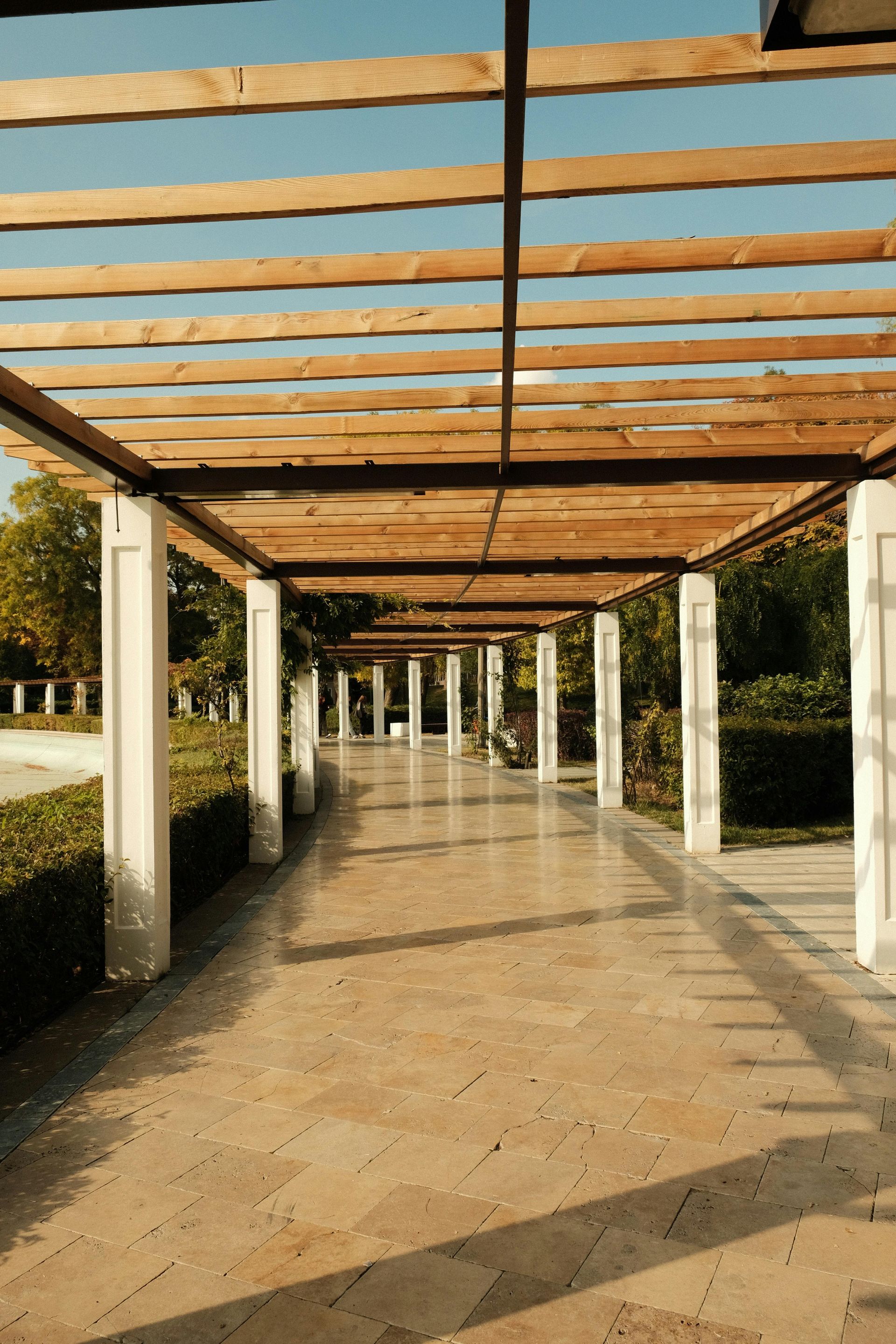 Stone pathway under a wooden pergola with white columns. Sunlight casts shadows.
