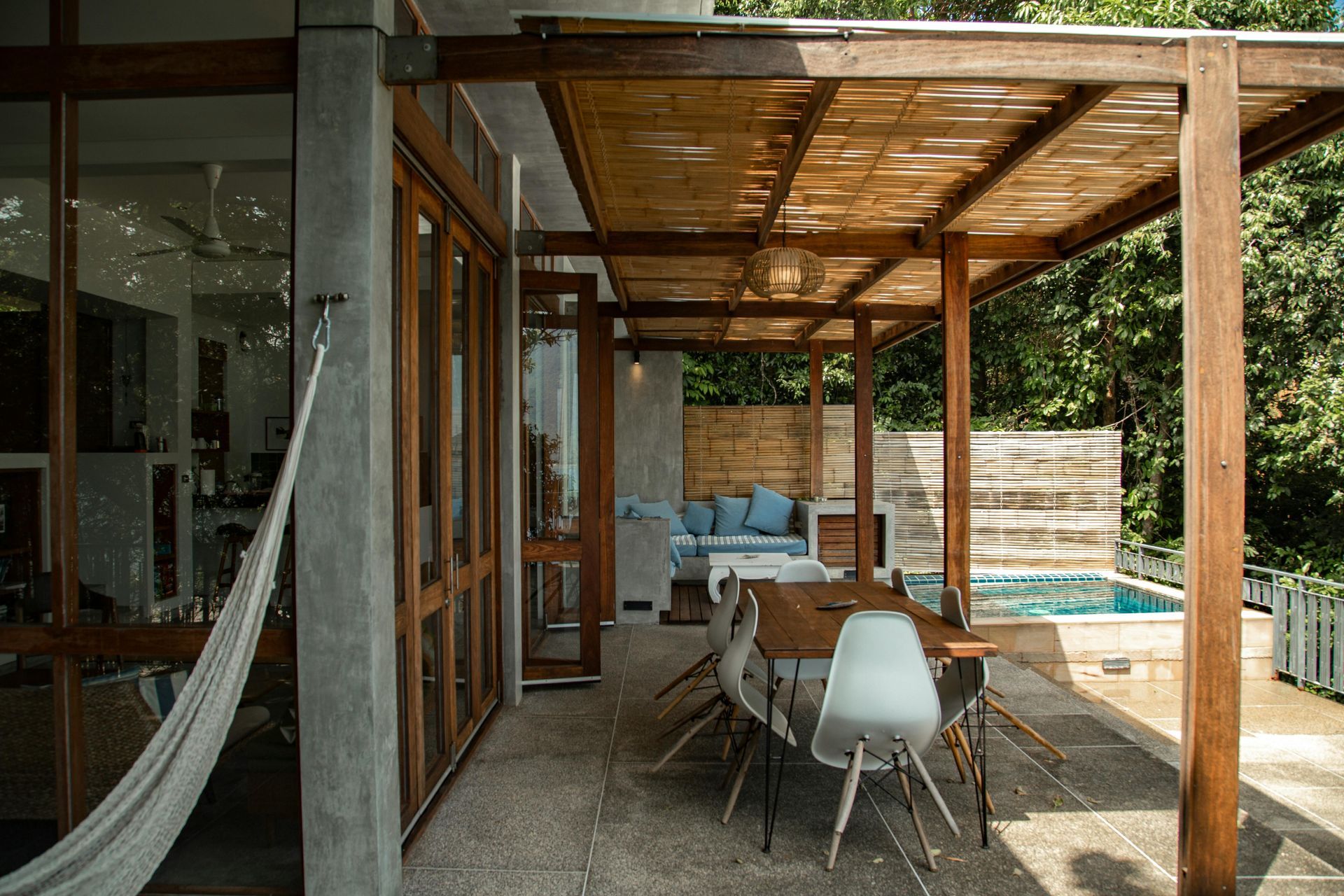 Patio with dining area, hammock, and pool, under a wooden pergola roof.