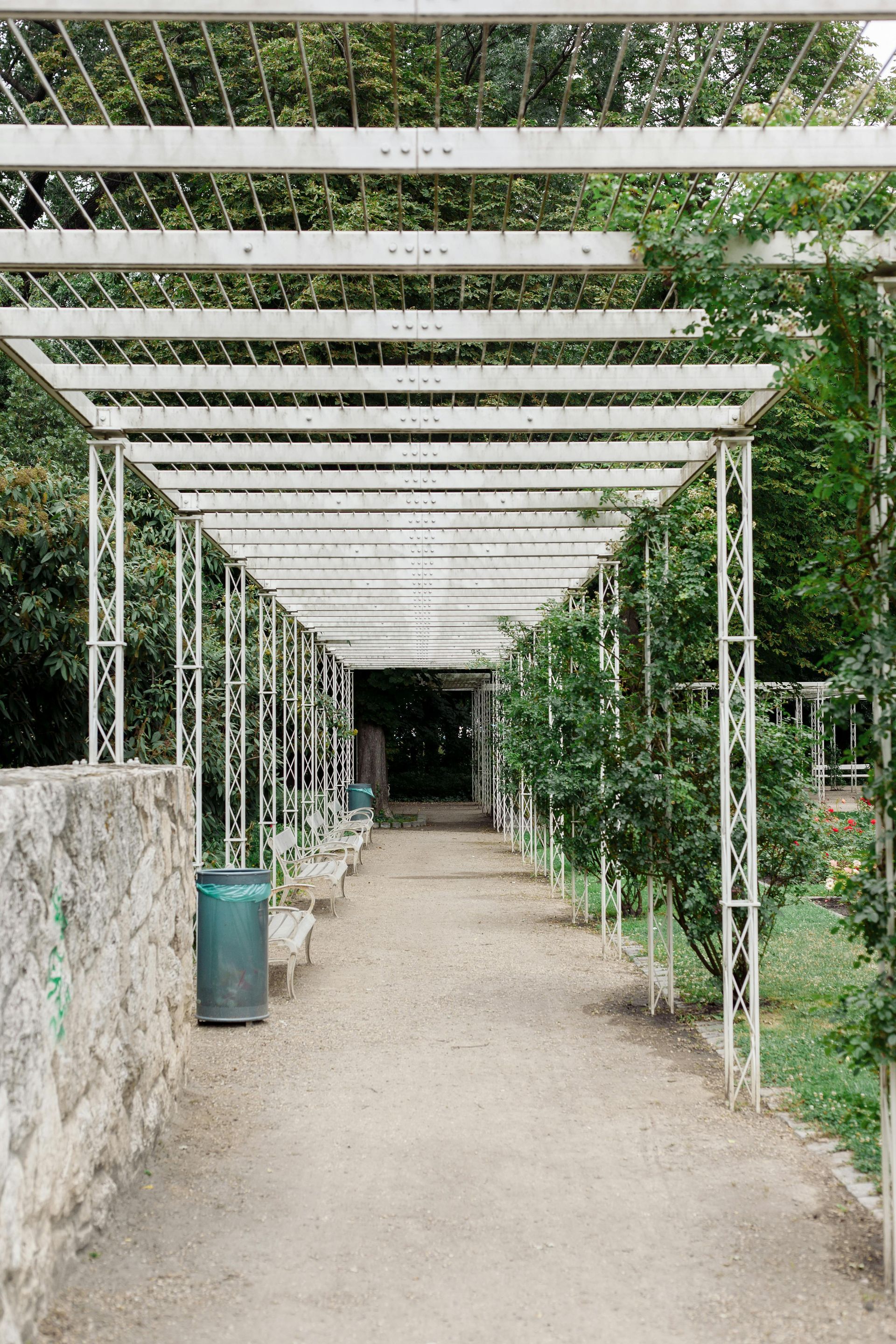 A gravel path leads through a white metal pergola with climbing plants, stone wall to the left.