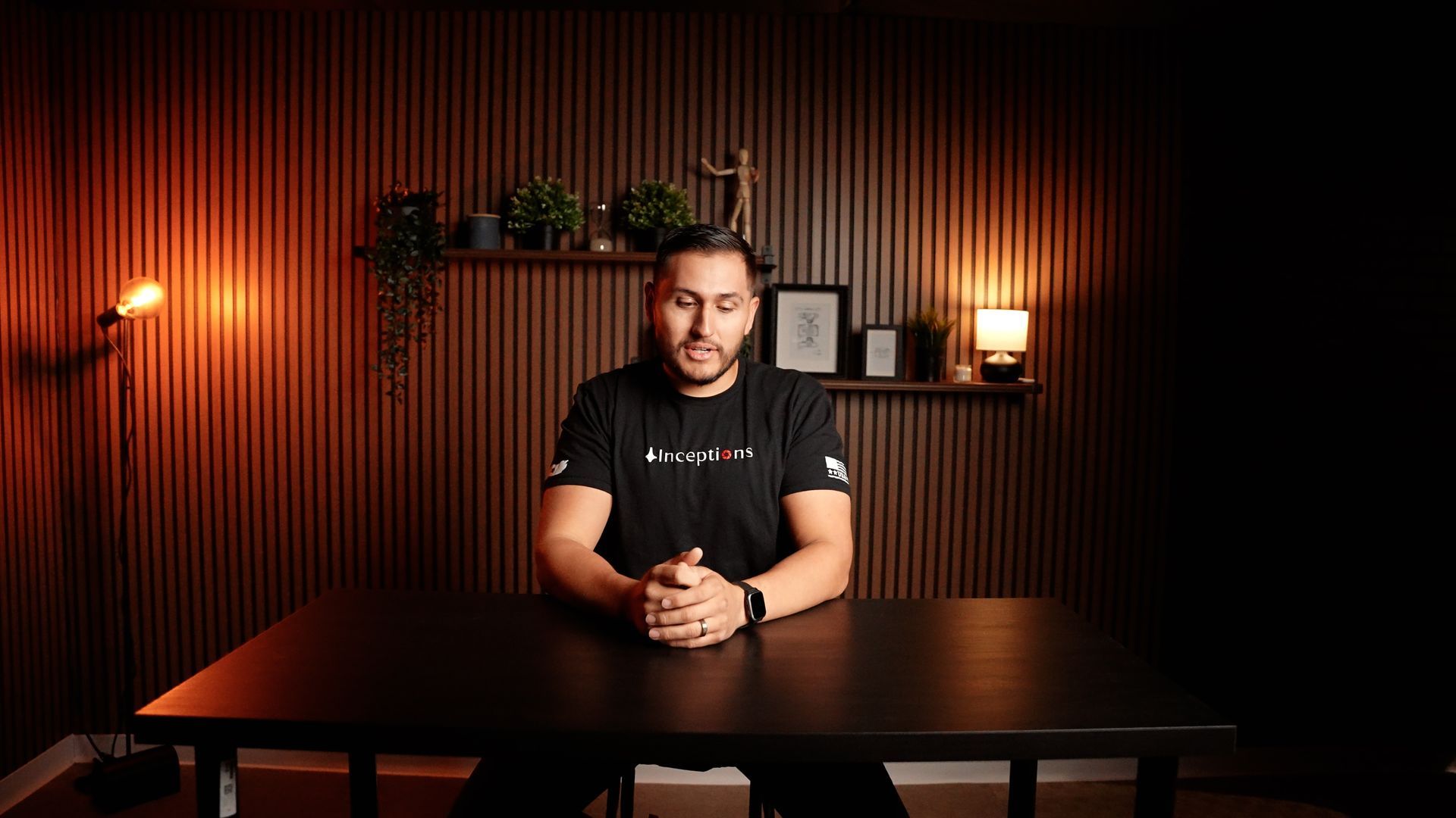 A man is sitting at a table in a dark room with his hands folded.