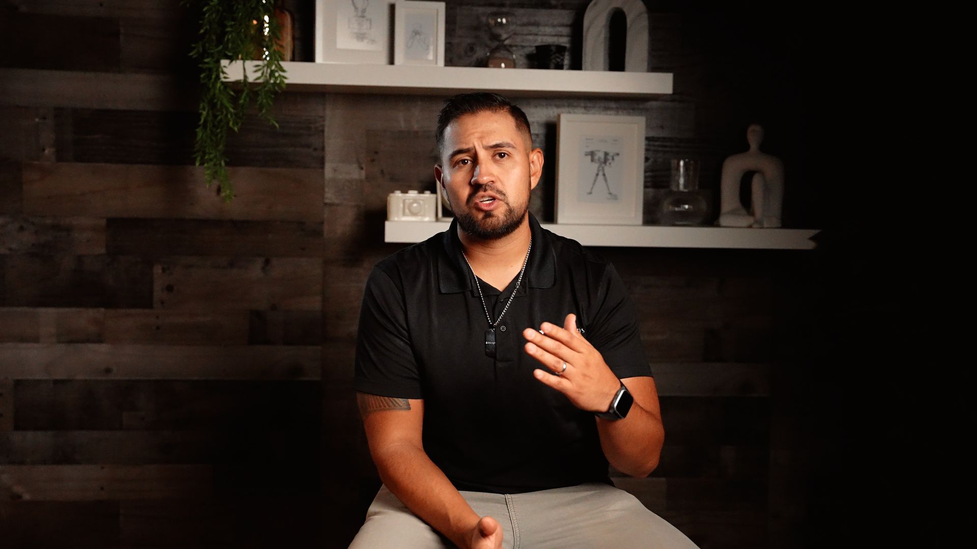 A man is sitting in front of a wooden wall and talking to the camera.