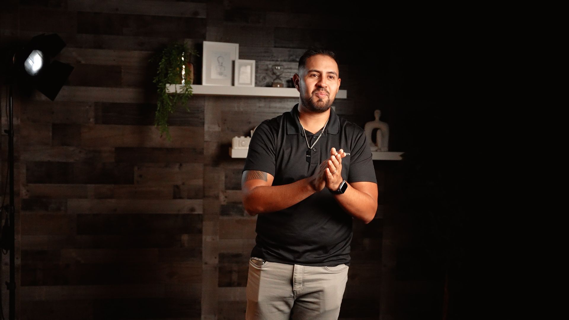 A man with a beard is standing in a dark room with his hands folded.
