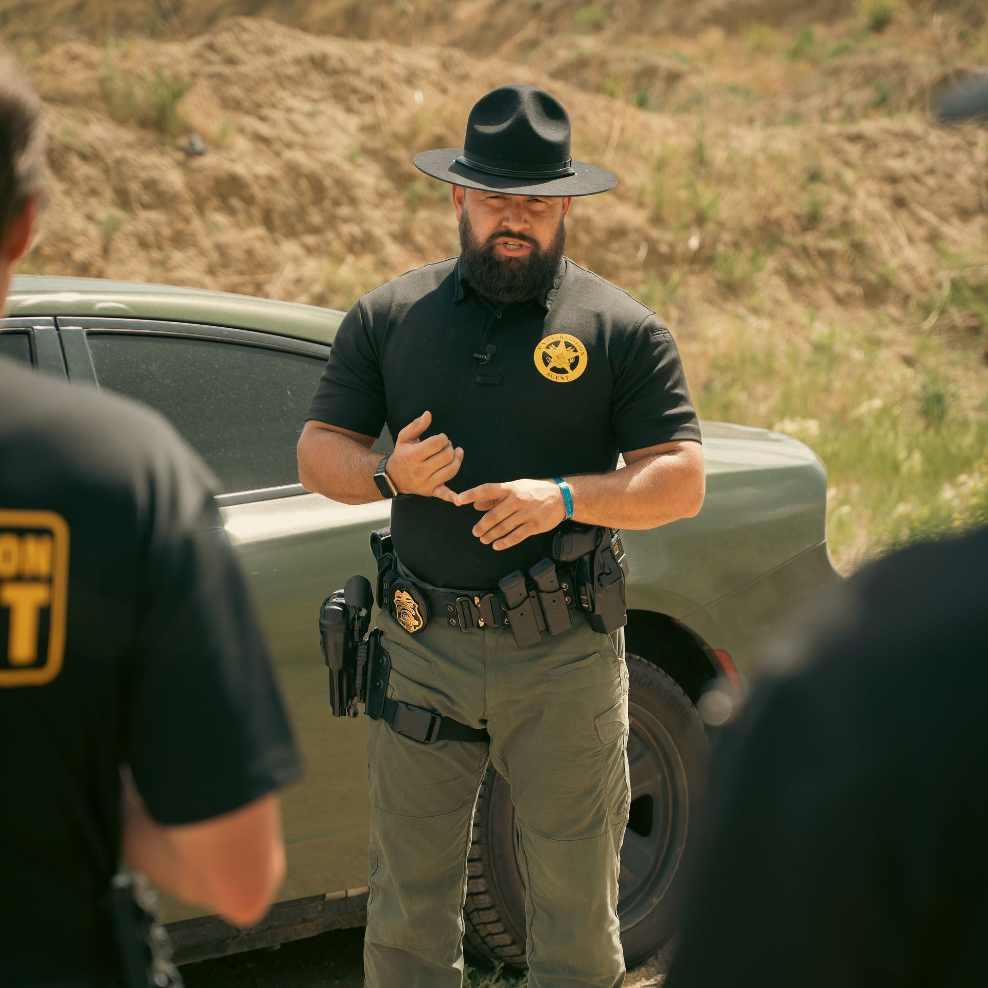 A bearded police officer wearing a hat stands in front of a car