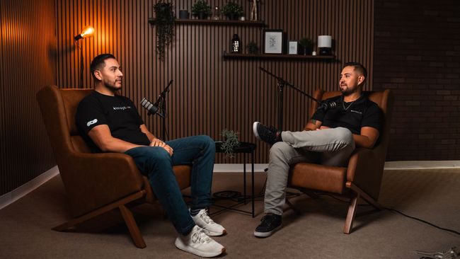 Two men are sitting in chairs in a room talking to each other.