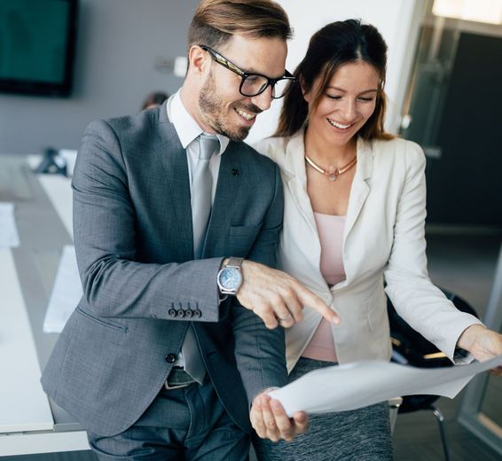 Man and woman in business attire smiling, pointing at a document indoors.