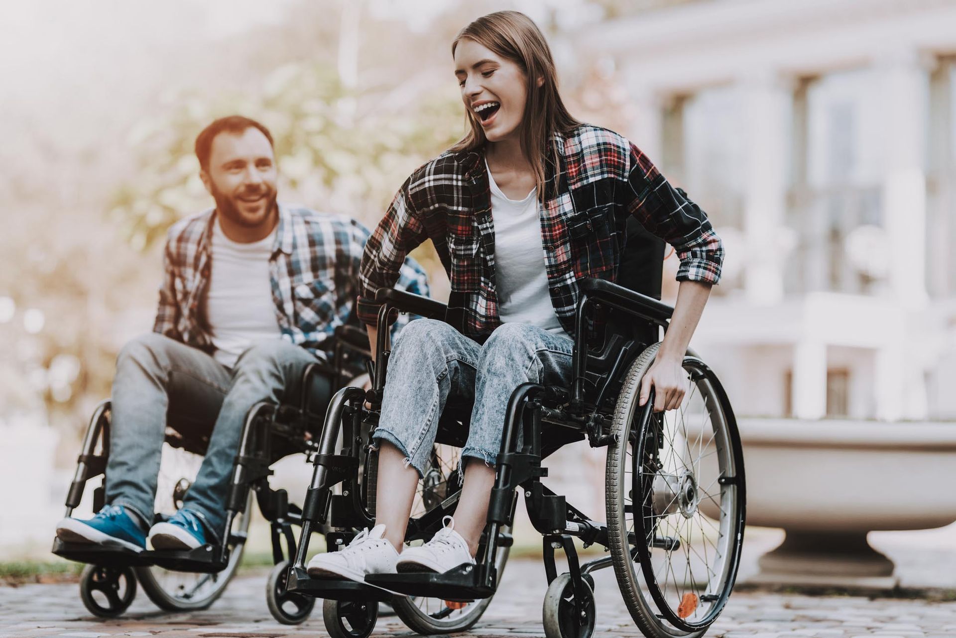 Two people in wheelchairs smile, outdoors. A woman laughs while the man looks at her.