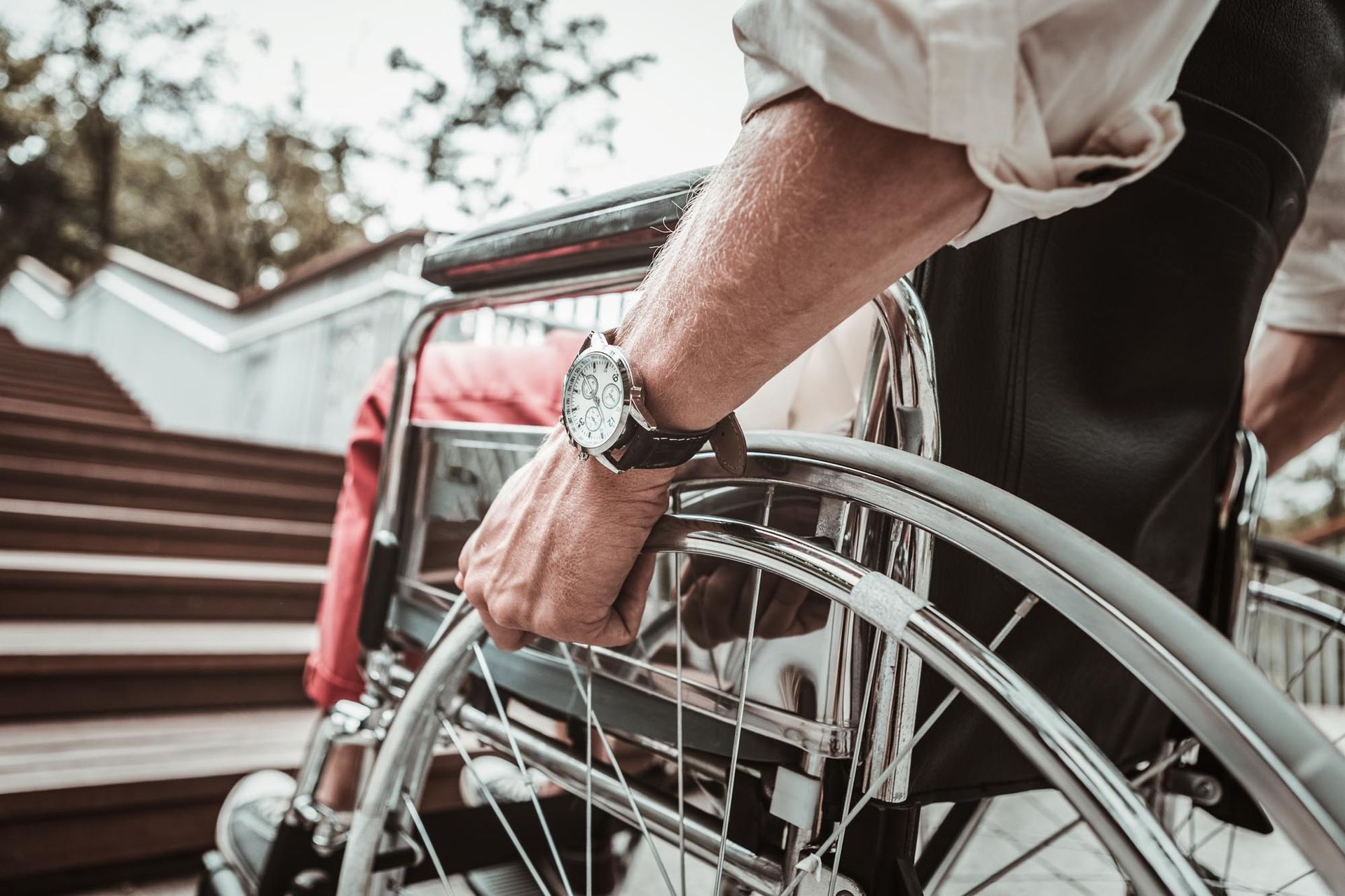 Man in a wheelchair, hand gripping wheel, near a set of stairs.