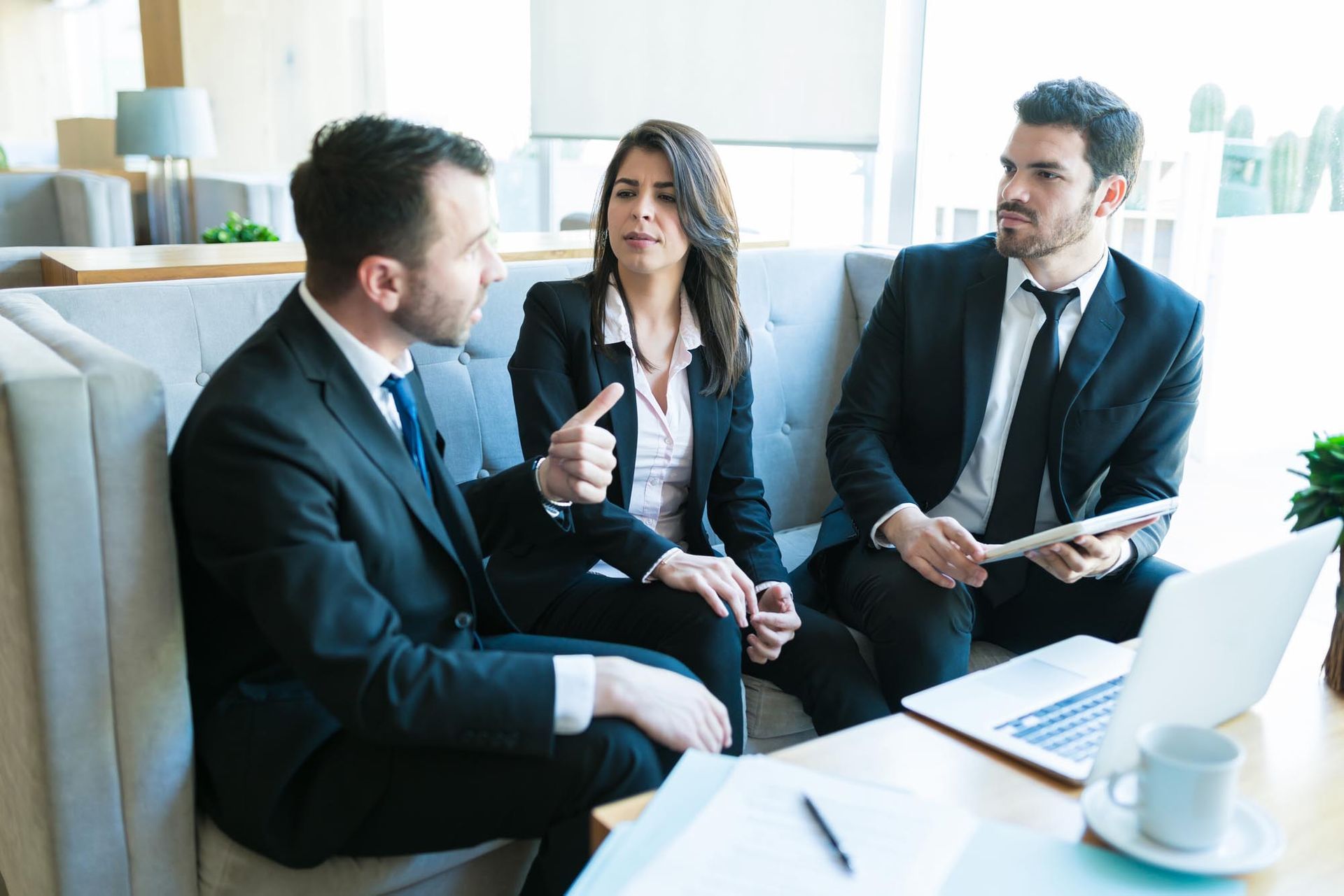 Three business people in suits discuss ideas while seated in a modern setting.