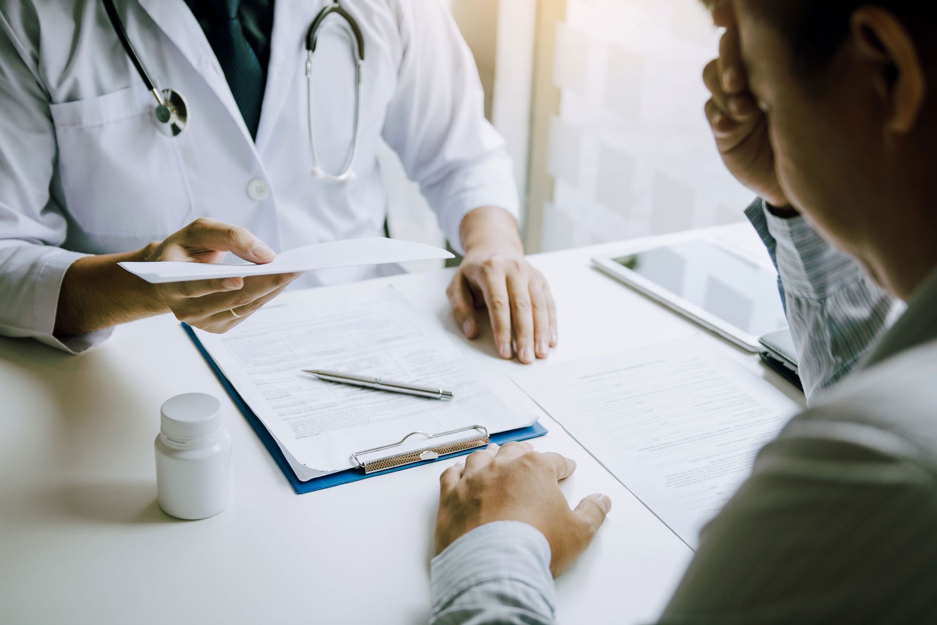 Doctor in white coat shows paperwork to a patient, who holds their head in their hand at an office desk.