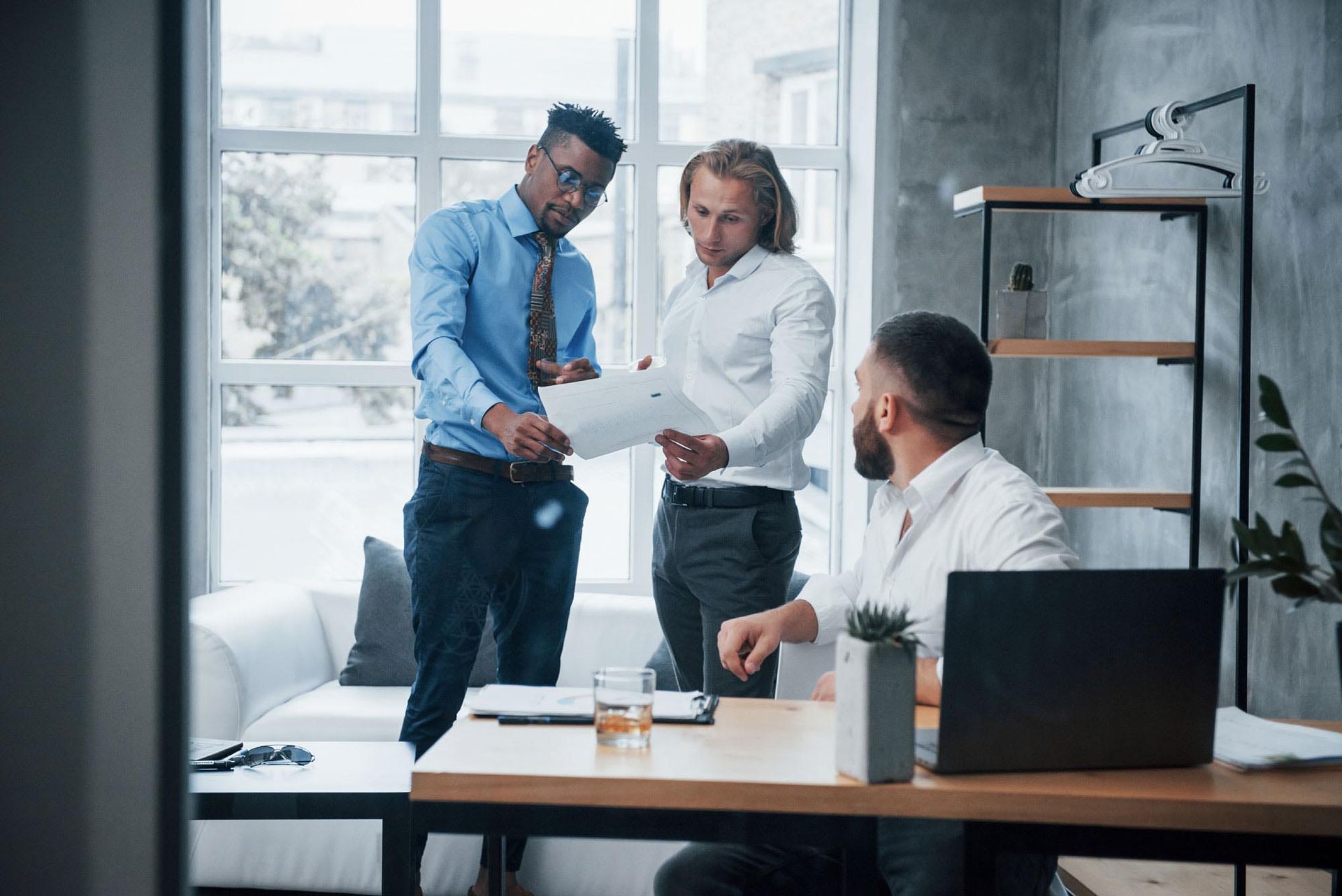 Three men in a modern office, reviewing documents. One points at paper, another looks on, the third at a desk with a laptop.