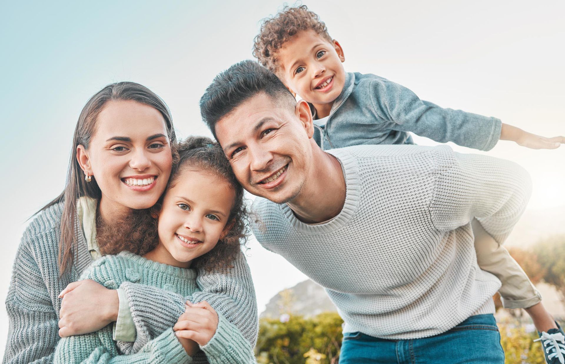 Smiling family of four posing outdoors. Mom embraces daughter; dad carries son on his back.