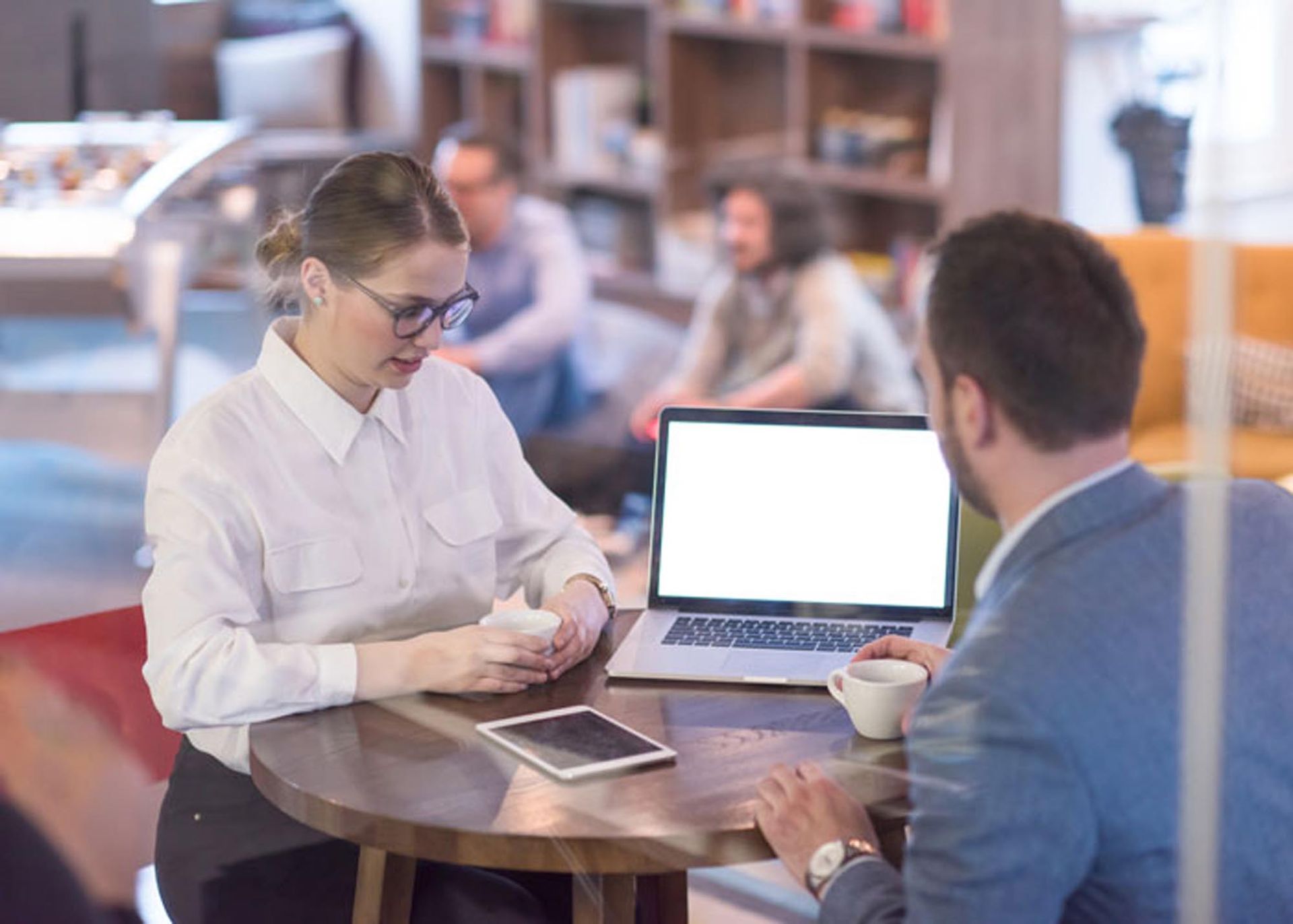 Woman and man in business attire at a table, laptop open, other people in background.
