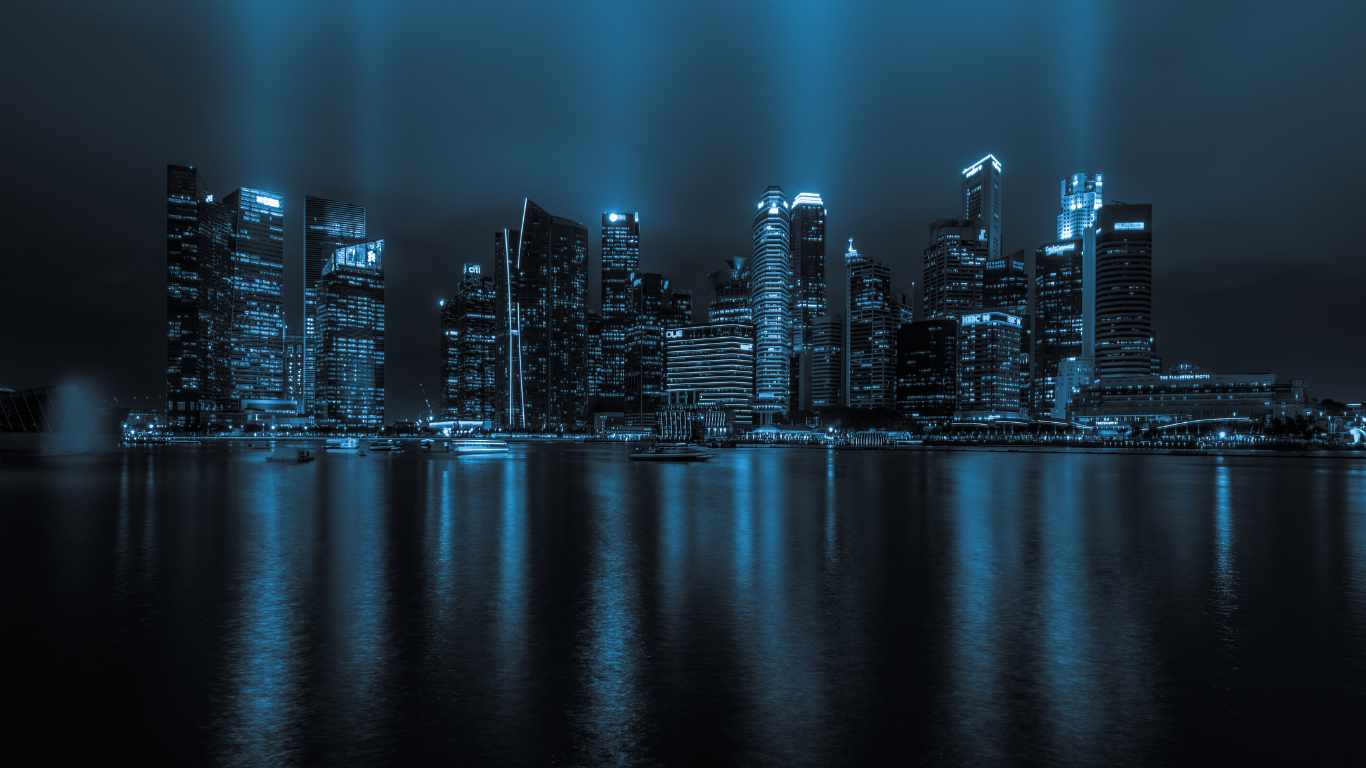Skyline notturno con fasci di luce sui grattacieli riflessi nell’acqua.