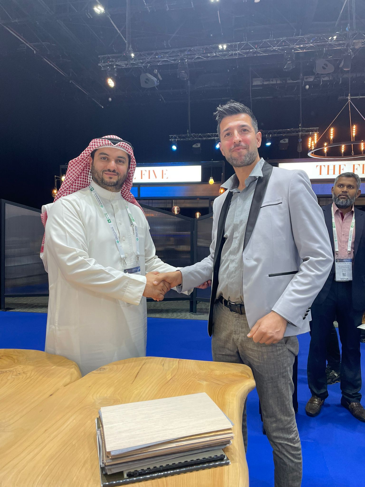 Due uomini si stringono la mano allo stand di un evento; uno indossa abiti tradizionali arabi, l'altro un abito.