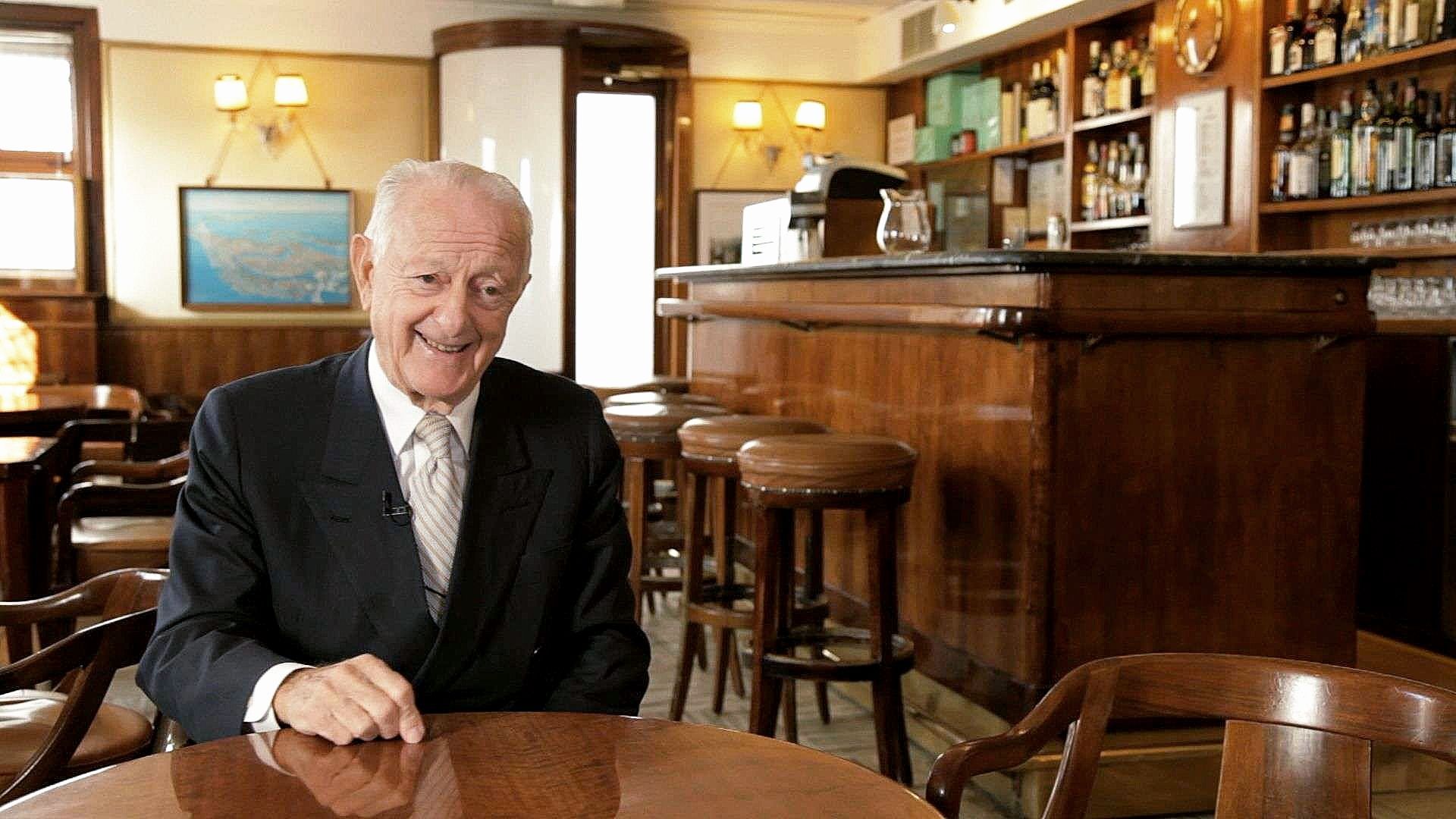 Un uomo in giacca e cravatta sorride seduto a un tavolo in un bar con pannelli in legno.