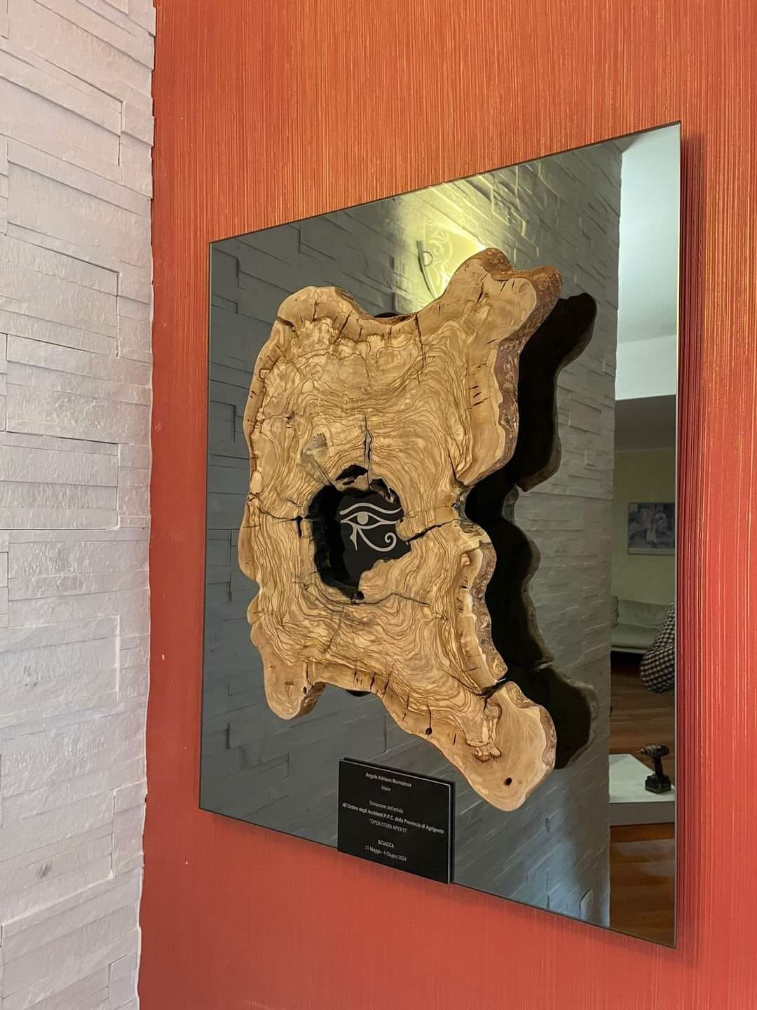 Opera d'arte montata a parete: una lastra di legno con una parte centrale scura su uno sfondo grigio di metallo e specchio, contro una parete rosso-arancio.