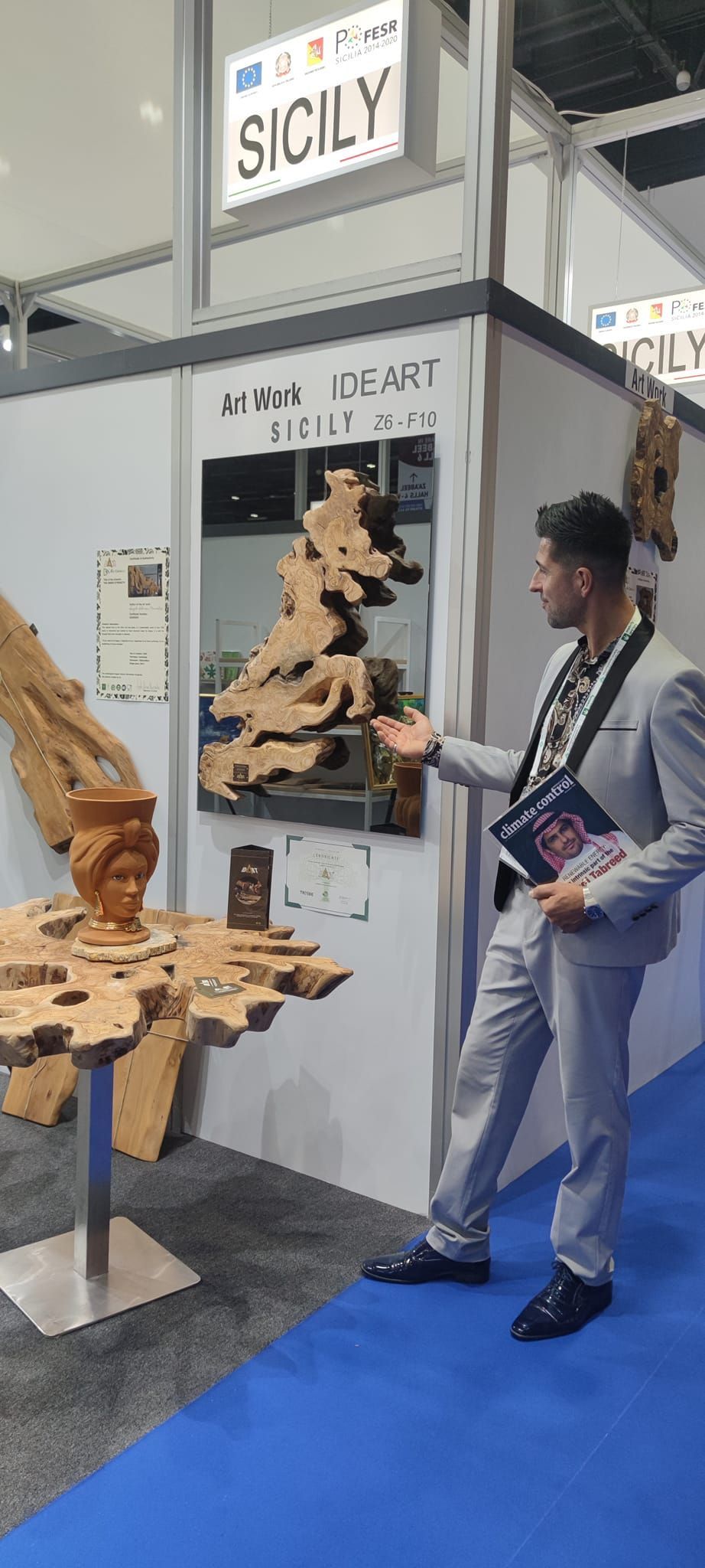 Uomo in giacca e cravatta alla mostra sulla Sicilia, indica un'opera d'arte. Moquette blu, pareti e cornice grigio chiaro.