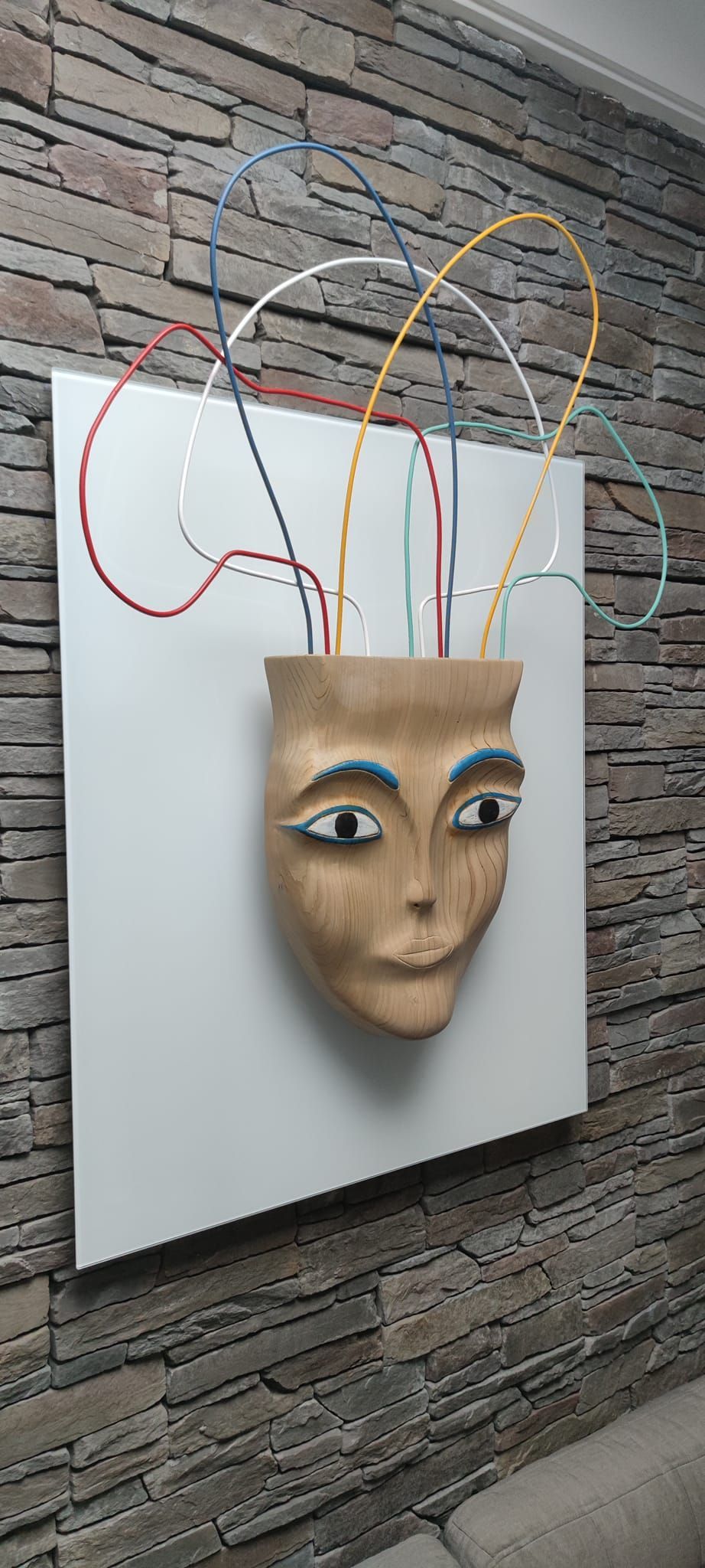 Un'opera d'arte murale raffigurante una mascherina con fili colorati che spuntano dalla parte superiore su un quadrato bianco.