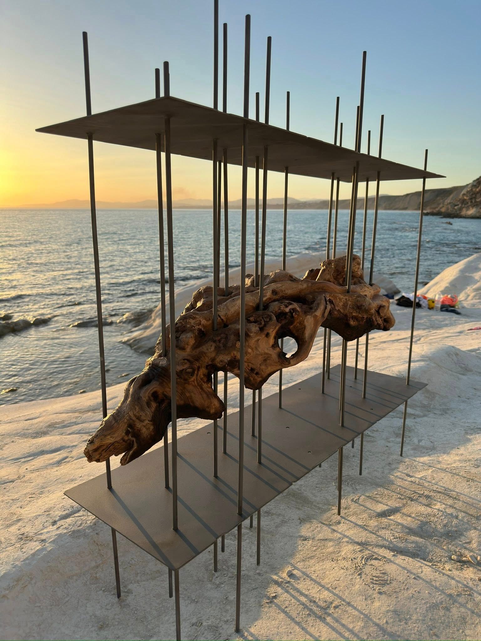 Scultura di legno galleggiante all'interno di una struttura metallica su una spiaggia, con tramonto sullo sfondo.