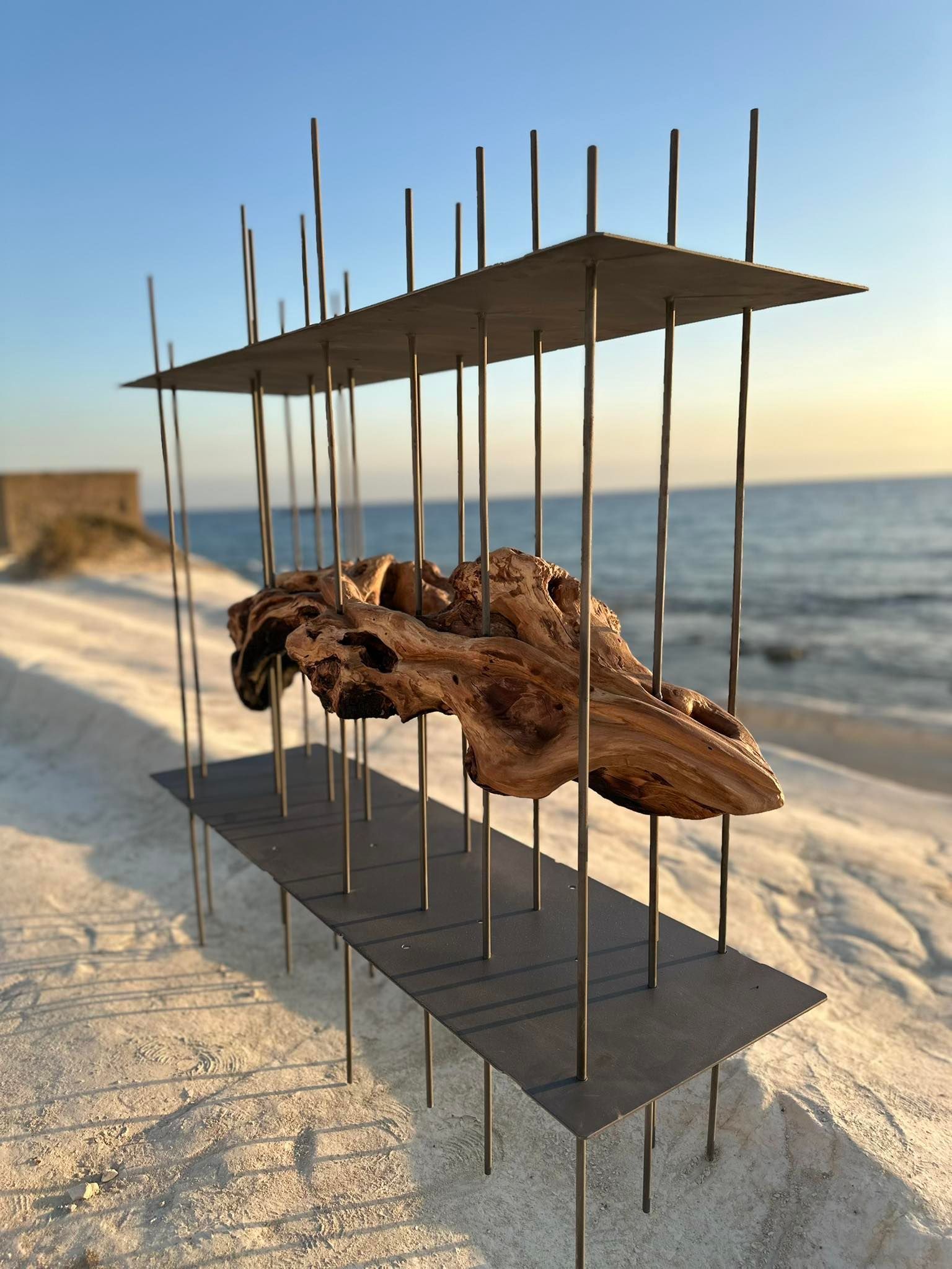 Scultura in legno galleggiante racchiusa da barre e piastre di metallo, su una spiaggia contro un cielo blu.