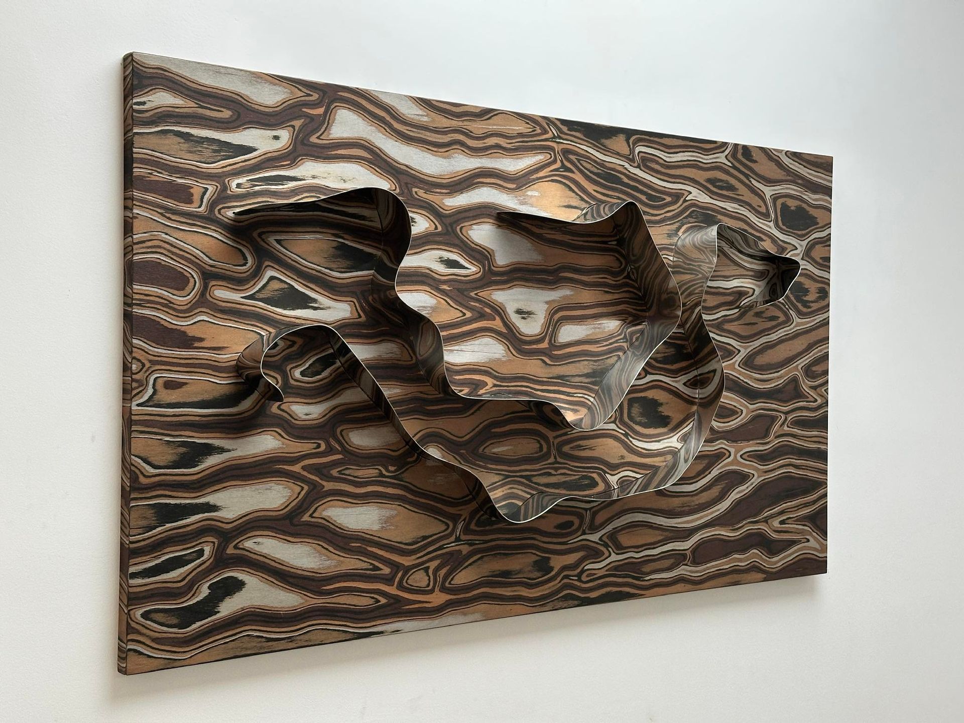 Scultura astratta in rilievo di metallo con motivi ondulati e forme ritagliate, montata su una parete bianca.