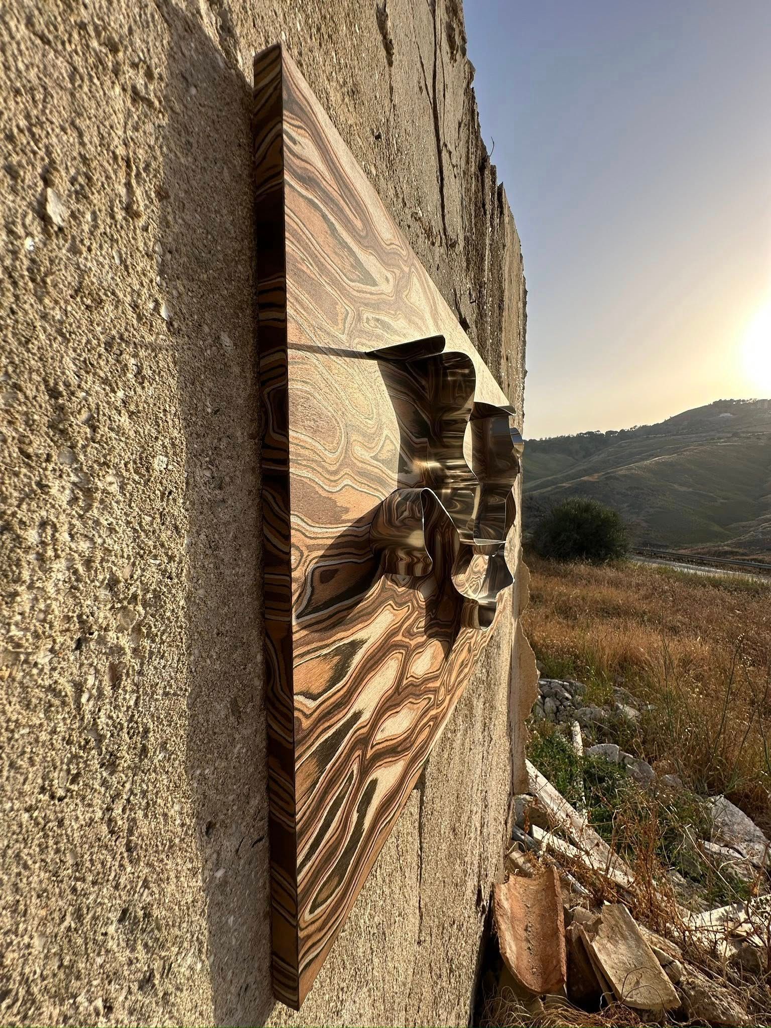 Opera d'arte in legno montata su un muro di pietra usurato; paesaggio sullo sfondo.