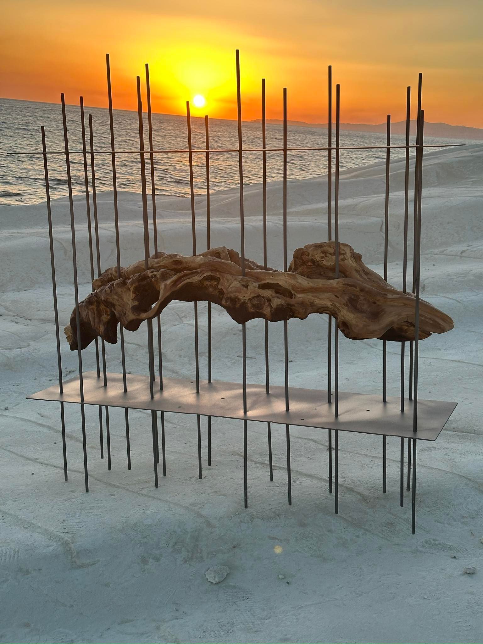 Scultura in legno marrone testurizzato, incorniciata da aste metalliche, posta su una superficie sabbiosa, sullo sfondo di un tramonto.