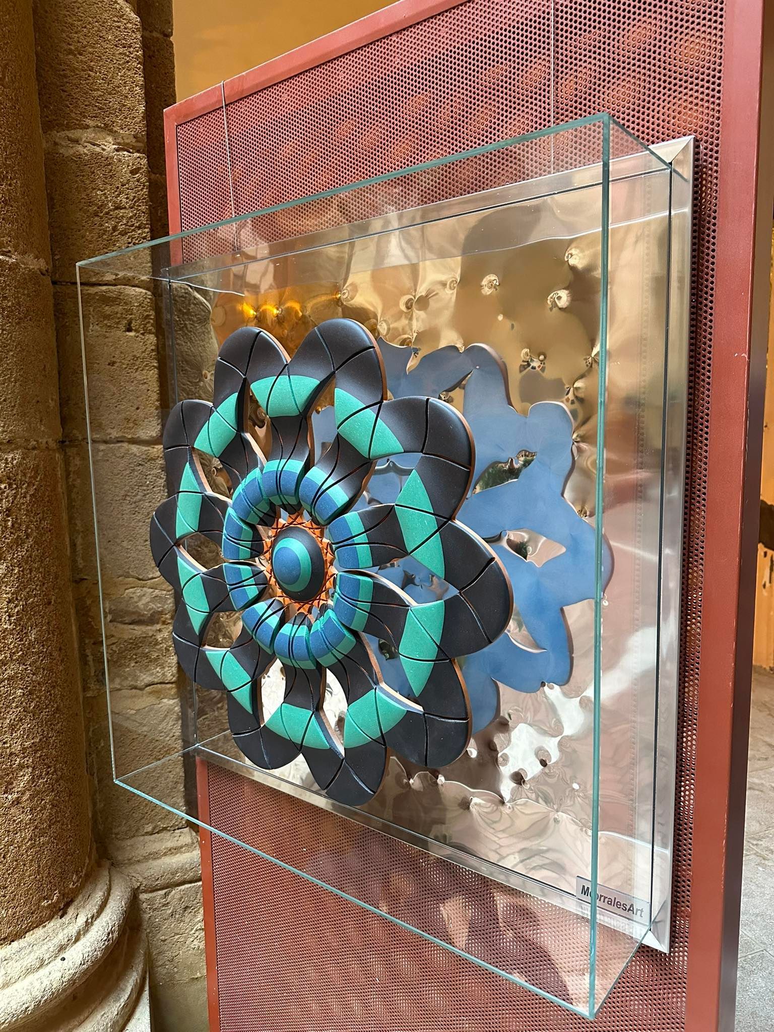 Opera decorativa in metallo in teca di vetro, con motivo floreale verde acqua e blu. Montata su un pannello marrone-rossastro.