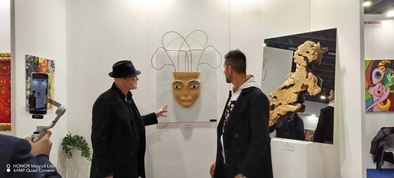 Due uomini osservano una maschera esposta a una mostra d'arte. Le pareti della mostra sono bianche e sono visibili altre opere d'arte.