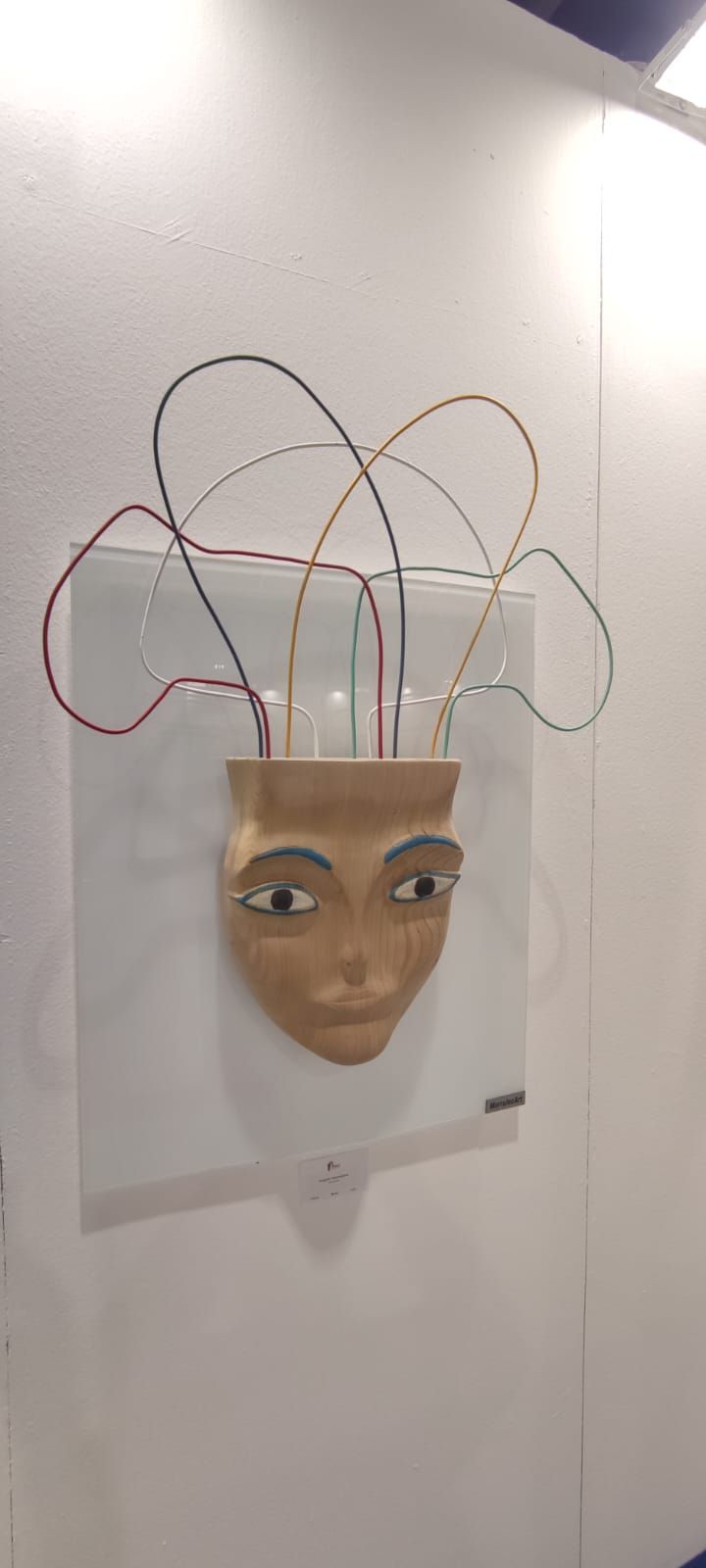 Una scultura da parete raffigurante un volto con fili che si estendono dalla parte superiore, posta su un quadrato bianco.